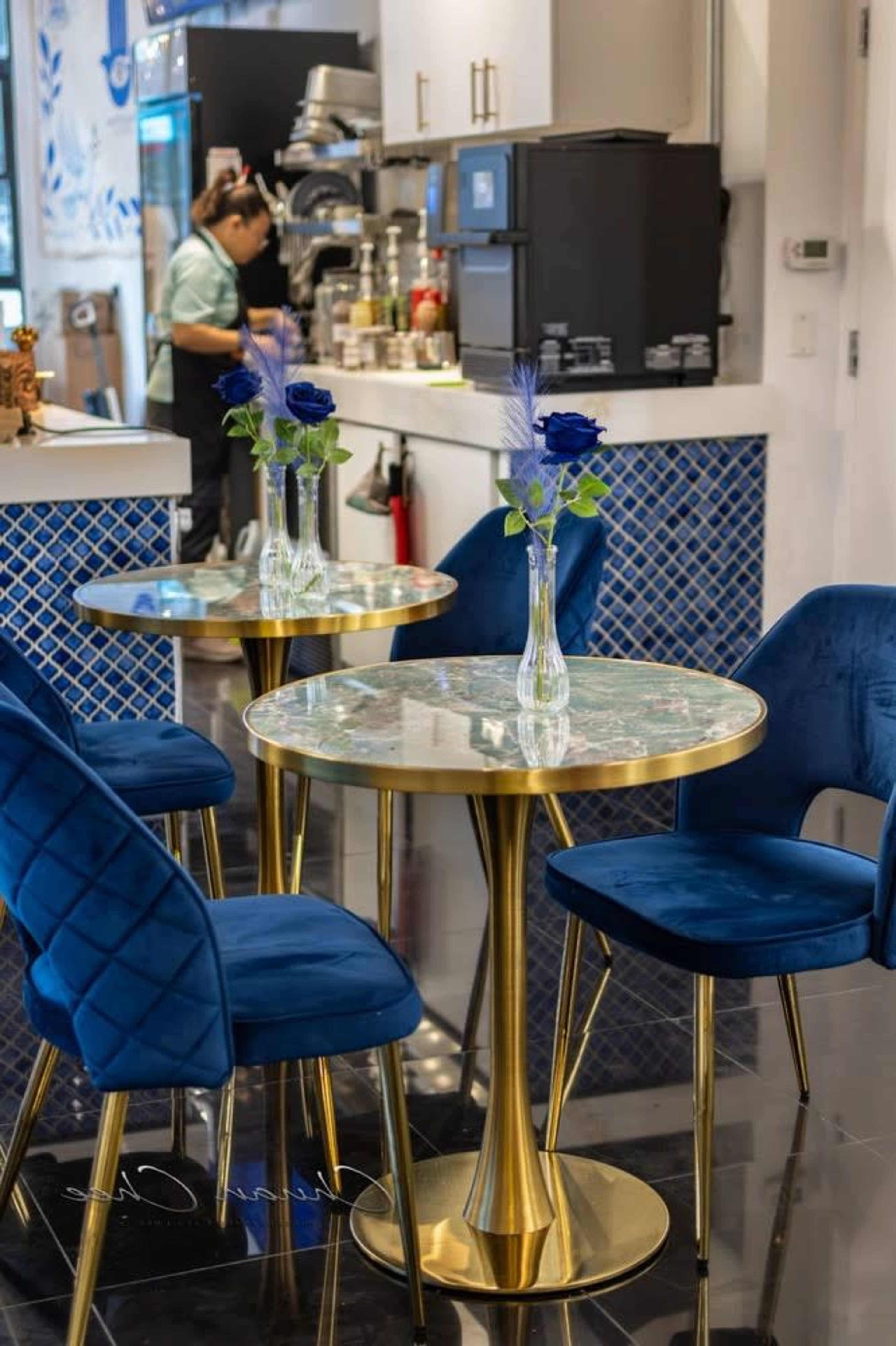 A small café features blue velvet chairs around round tables with gold bases, adorned with vases holding roses, and a coffee station in the background.