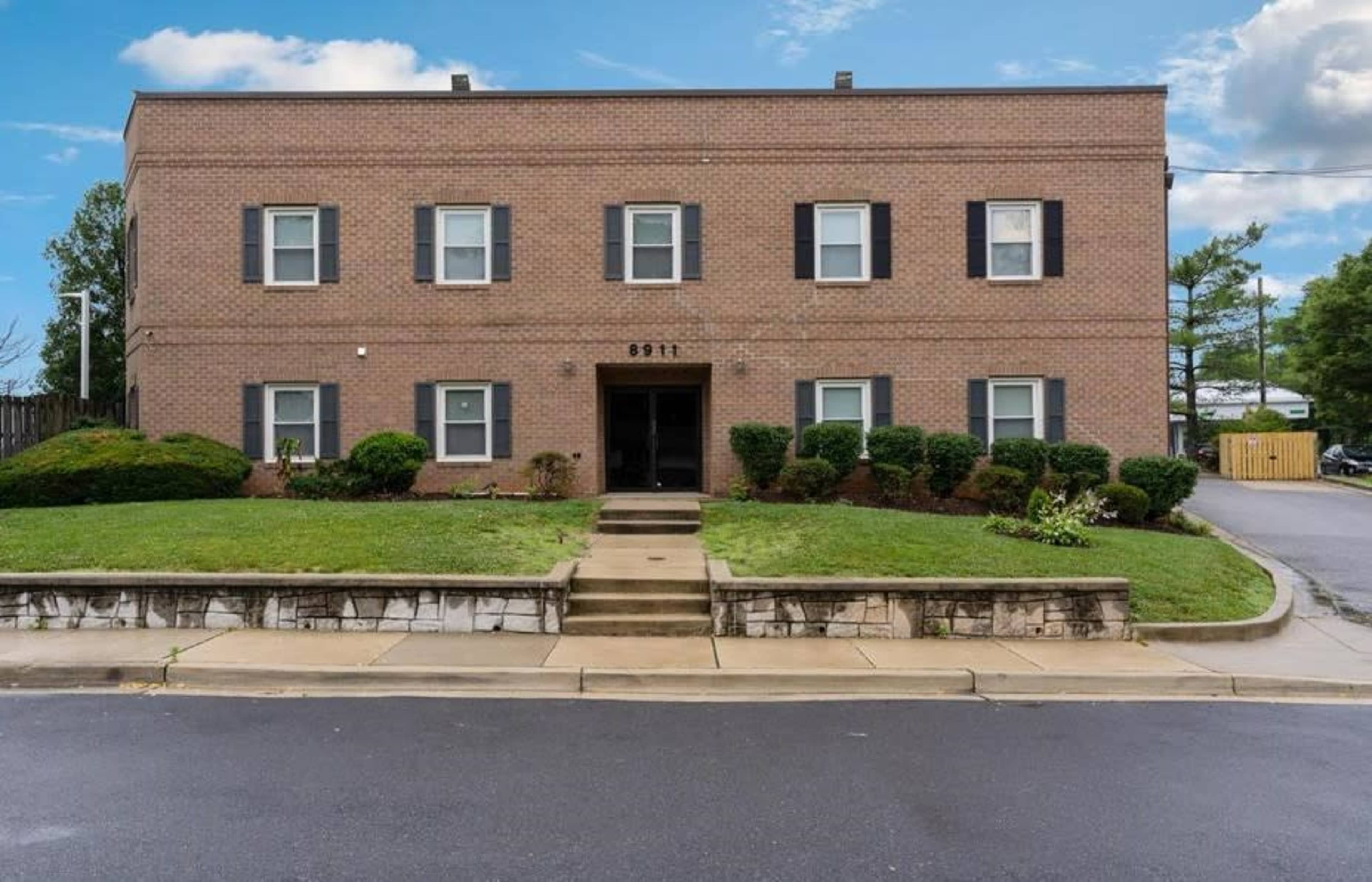 A two-story brick apartment building with black shutters and neatly trimmed bushes is located on a landscaped front lawn.