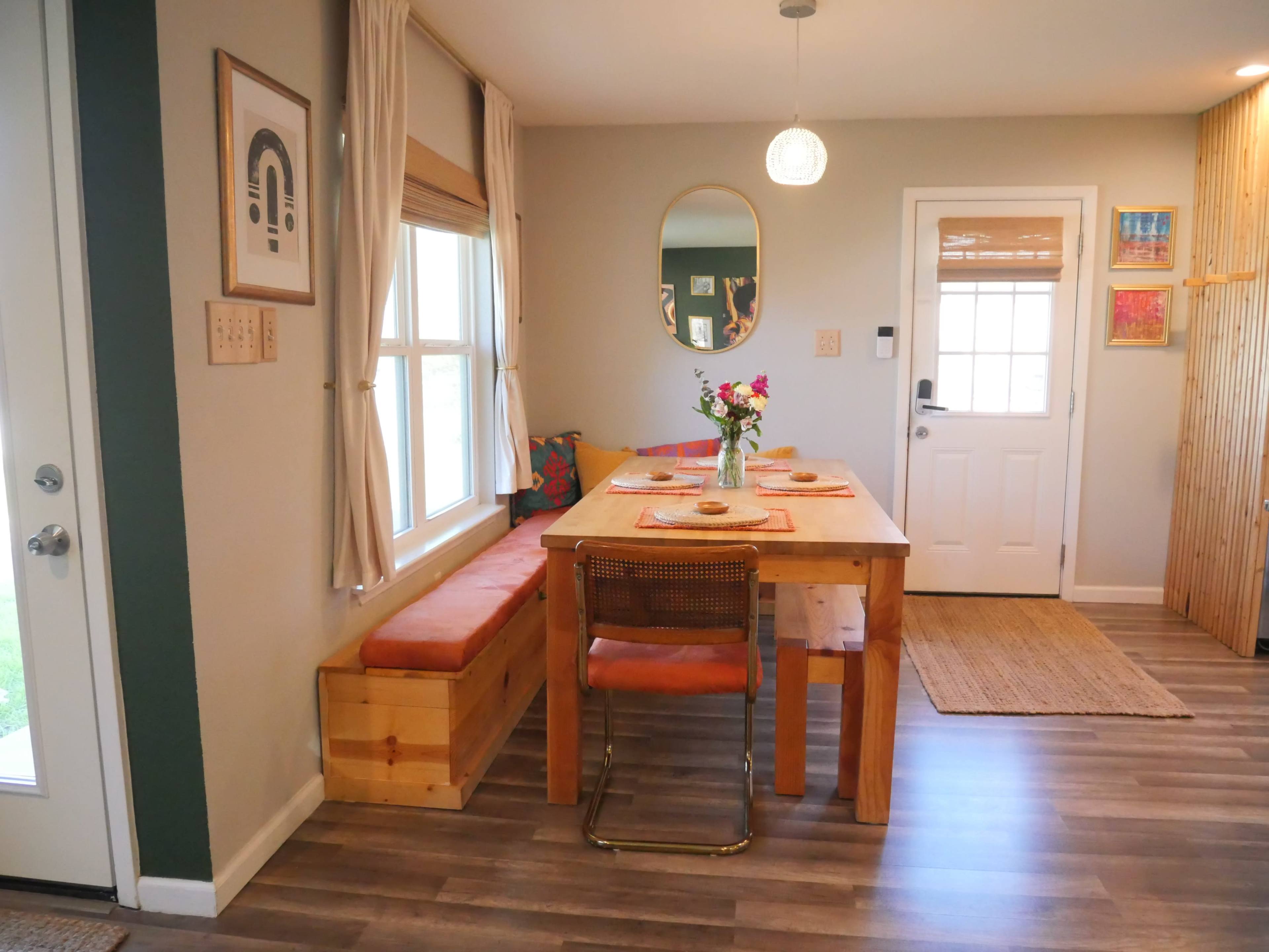 A wooden dining table with seating and a floral centerpiece is set up in a well-lit room next to a window, with a door leading outside in the background.