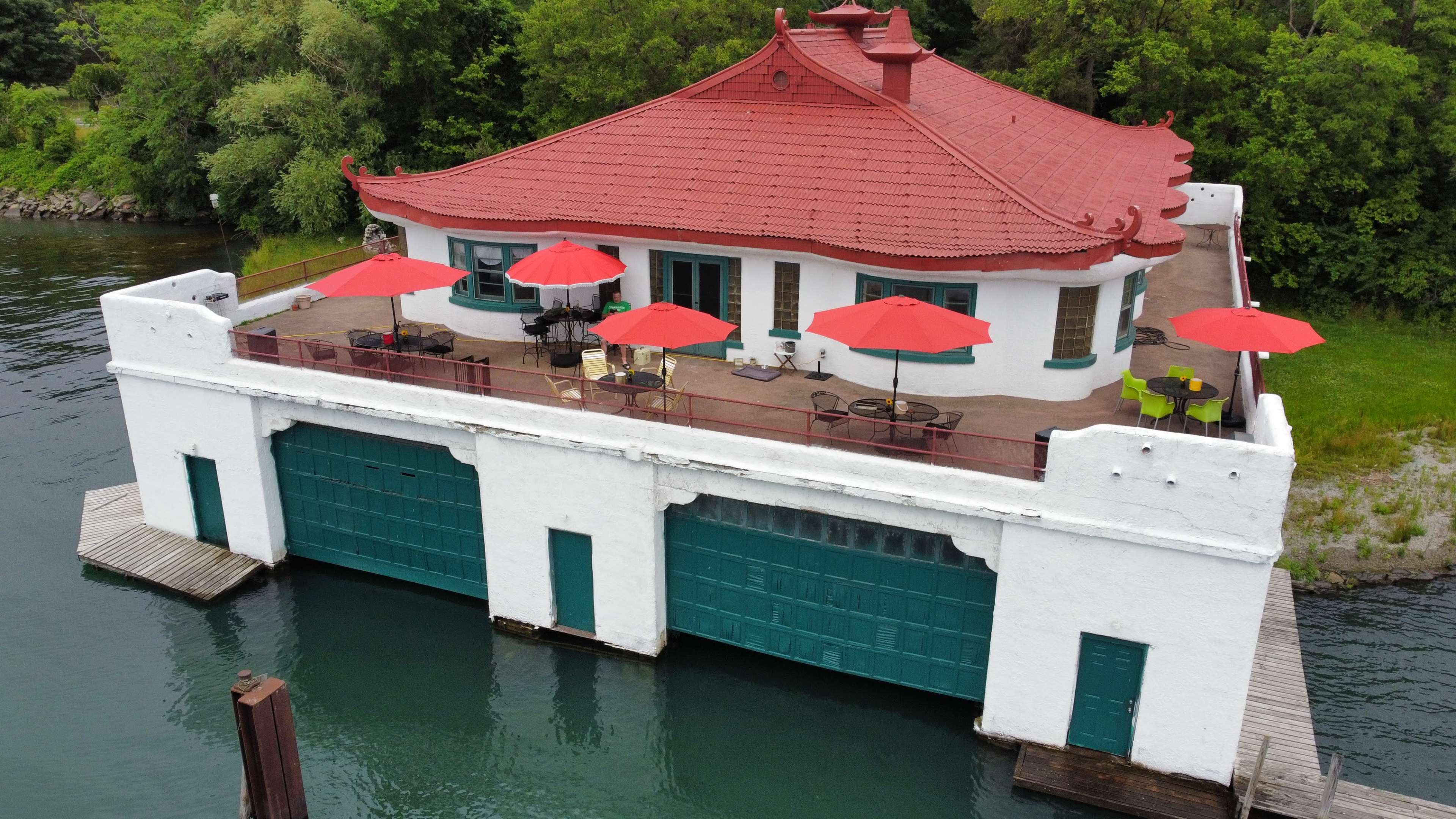 A white building with a red-tiled roof features a large deck with red umbrellas, situated above the water next to two green garage doors.