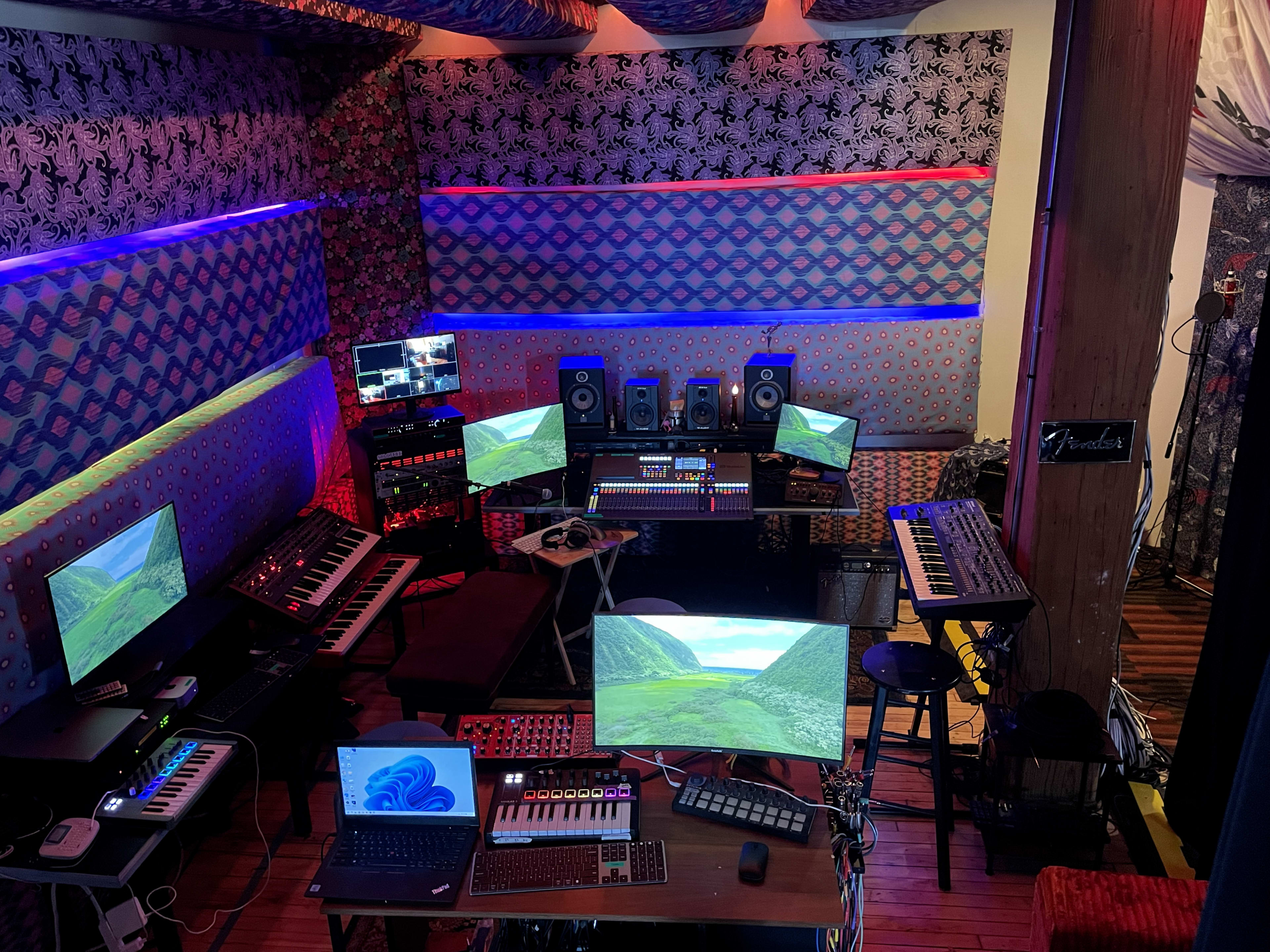 The image shows a music studio featuring multiple computers, synthesizers, and audio equipment arranged in a vibrant space with colorful wall designs and lighting.
