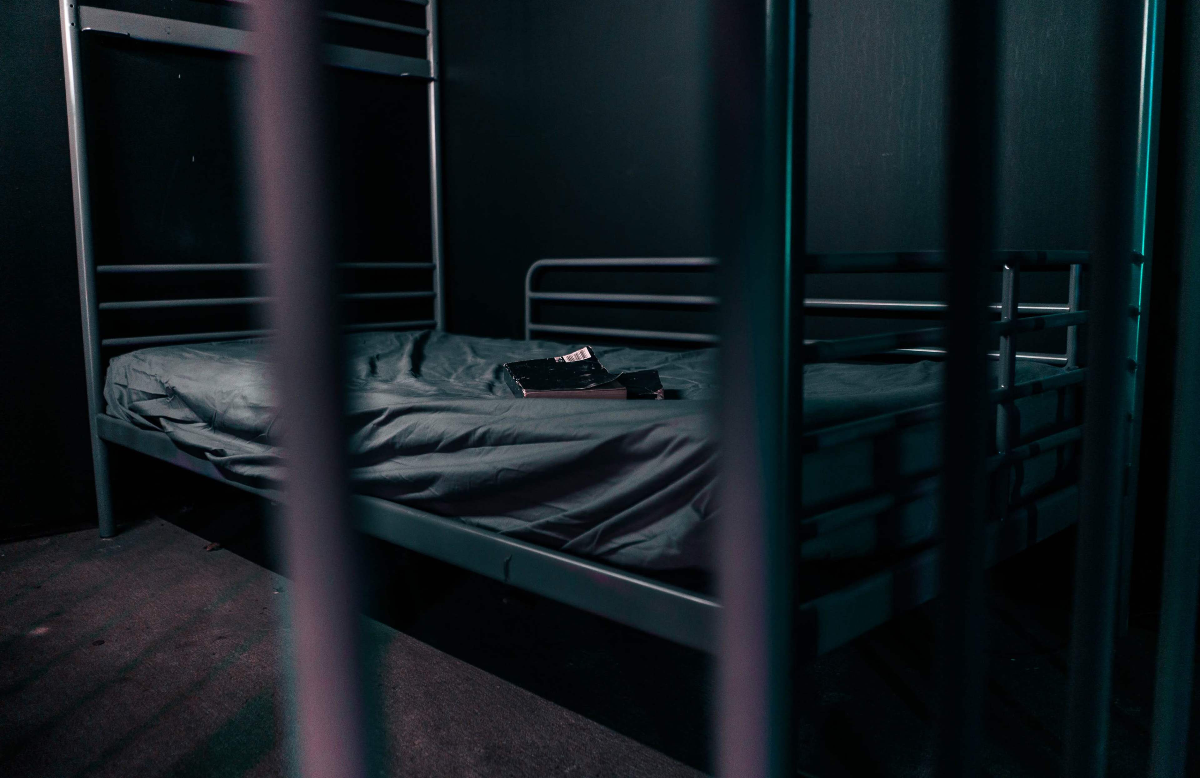 A metal-framed bed is positioned in a dimly lit room, with a rumpled mattress and a few items placed on top.