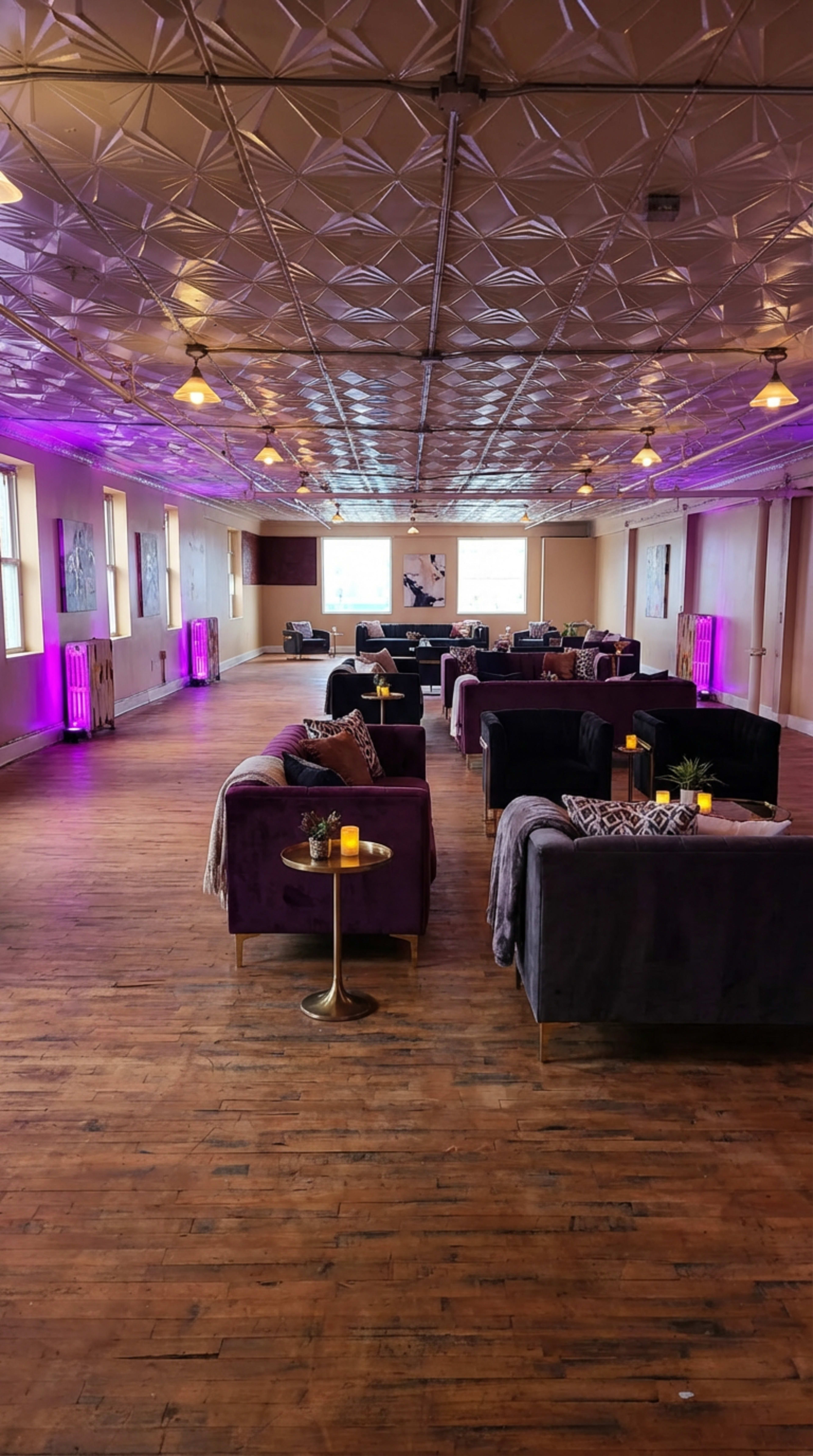 The image shows a spacious room with wooden floors, arranged with several velvet couches and tables, illuminated by soft purple lights.