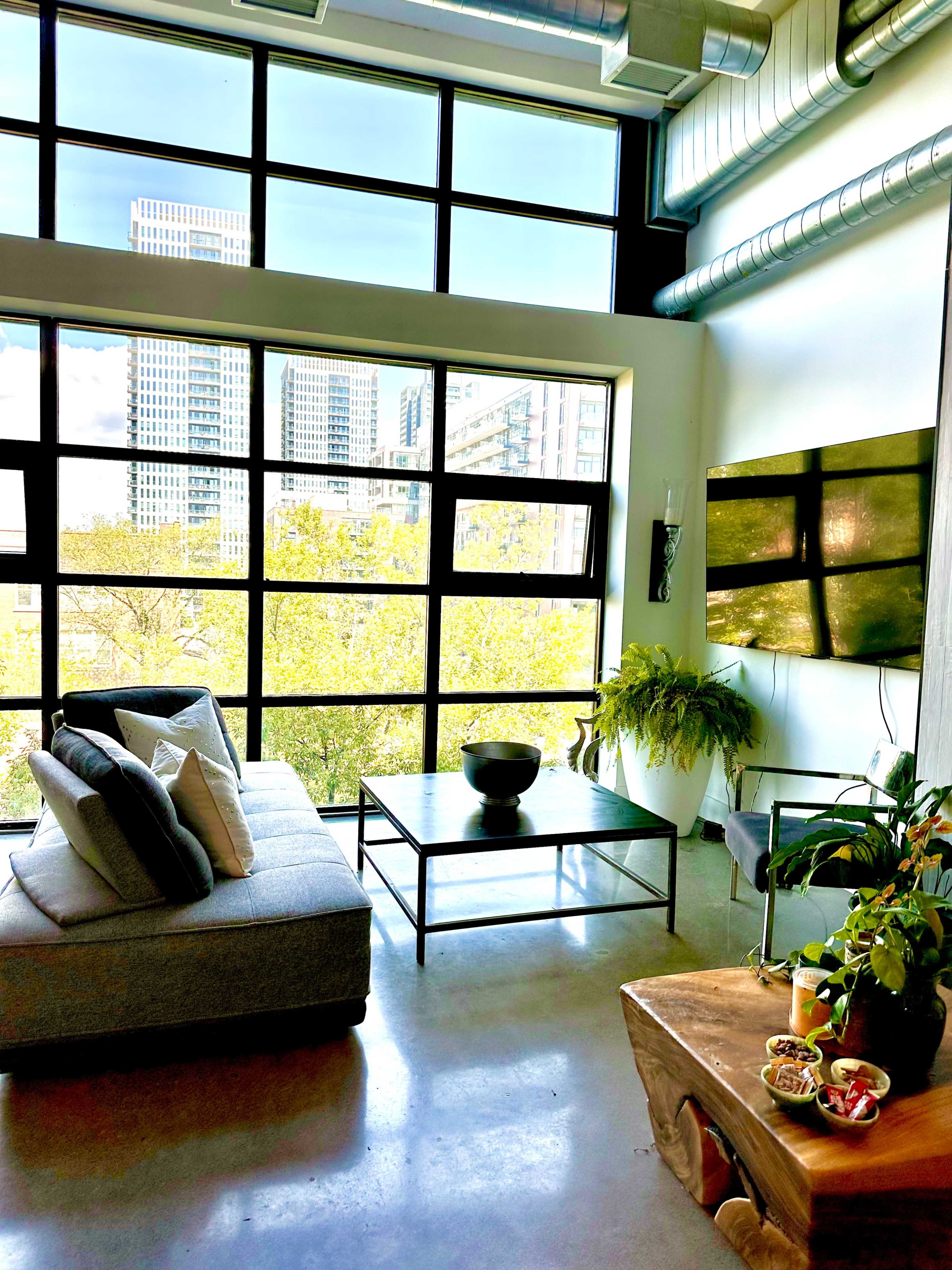 The image shows a modern living room with large windows overlooking a cityscape, featuring a gray sofa, a coffee table, and a decorative bowl on a nearby table.