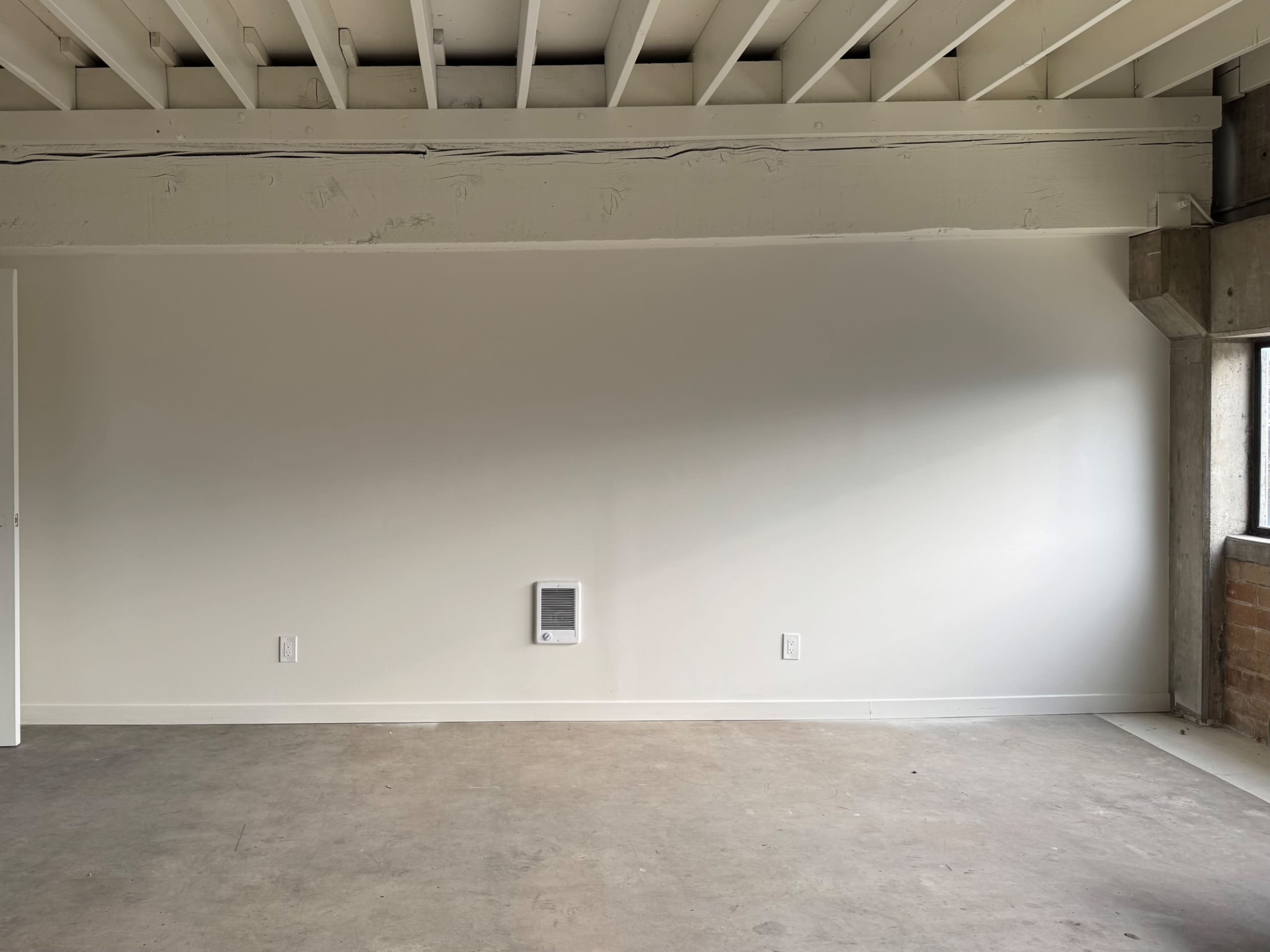 The image shows an empty room with a concrete floor, a plain white wall, and a small wall-mounted heater.