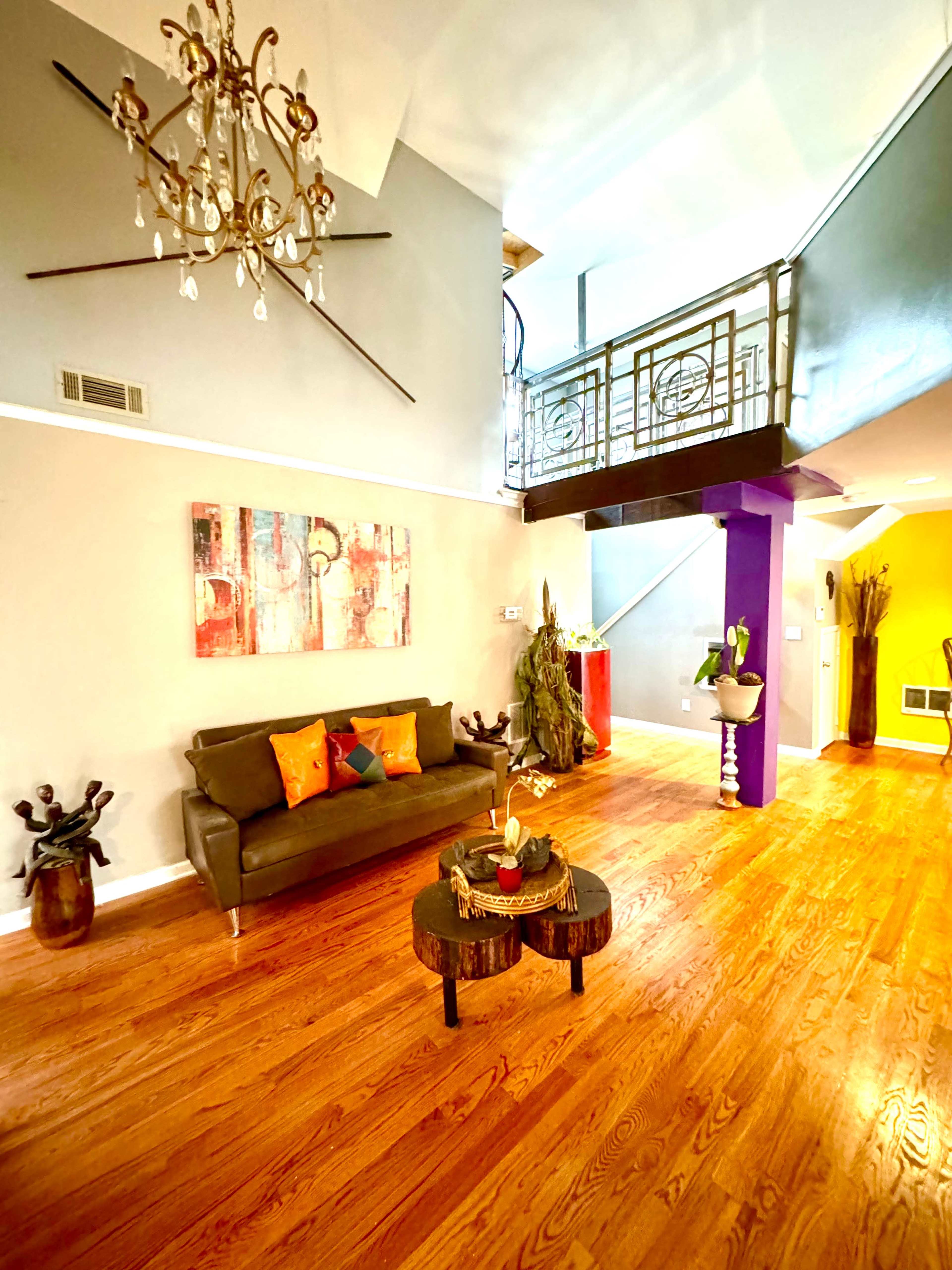 A living room with a dark sofa, colorful cushions, a round coffee table, and a tall plant, set against a backdrop of hardwood flooring and a staircase with a decorative railing.