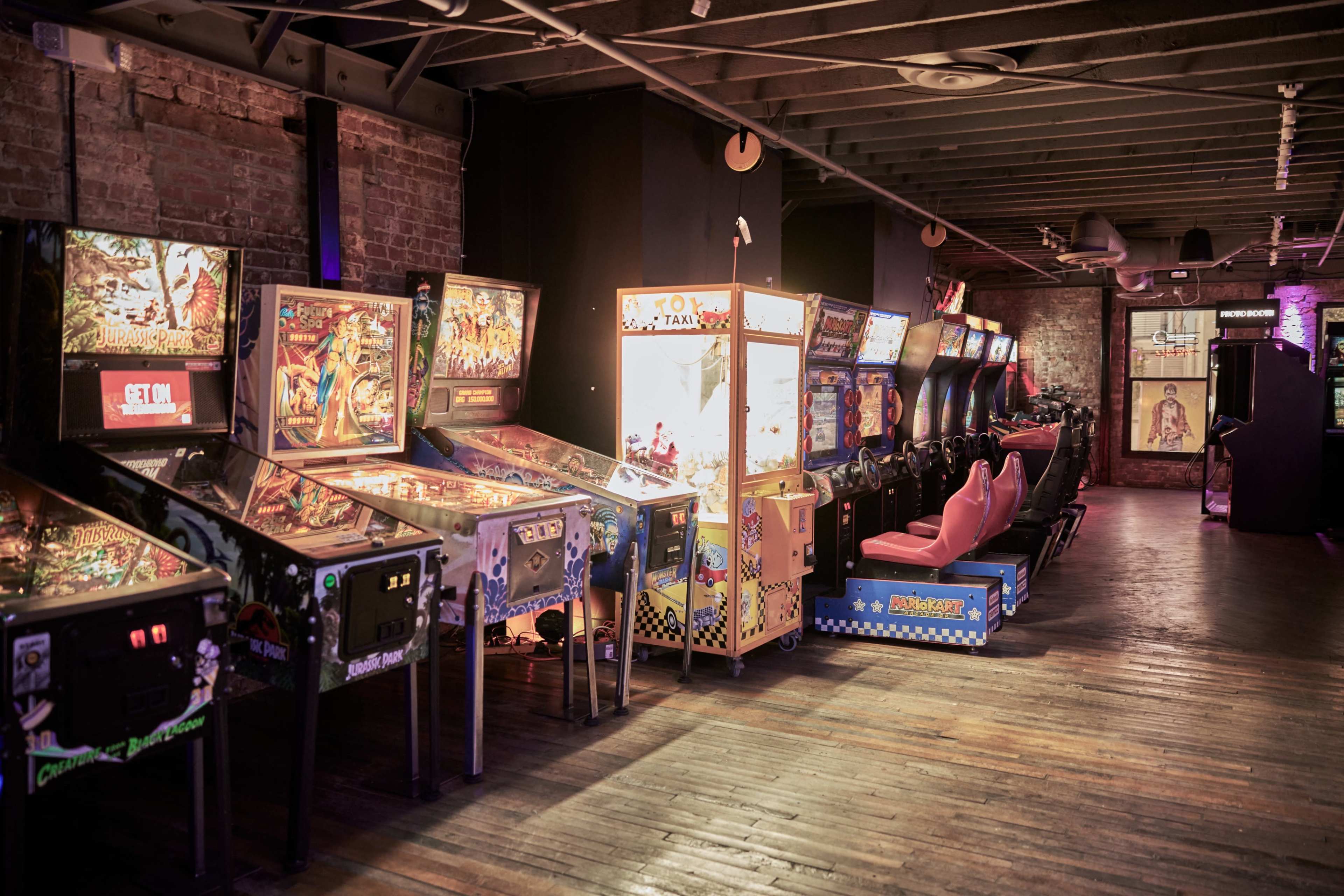 A row of vintage arcade machines and pinball tables in a dimly lit room with brick walls and wooden flooring.