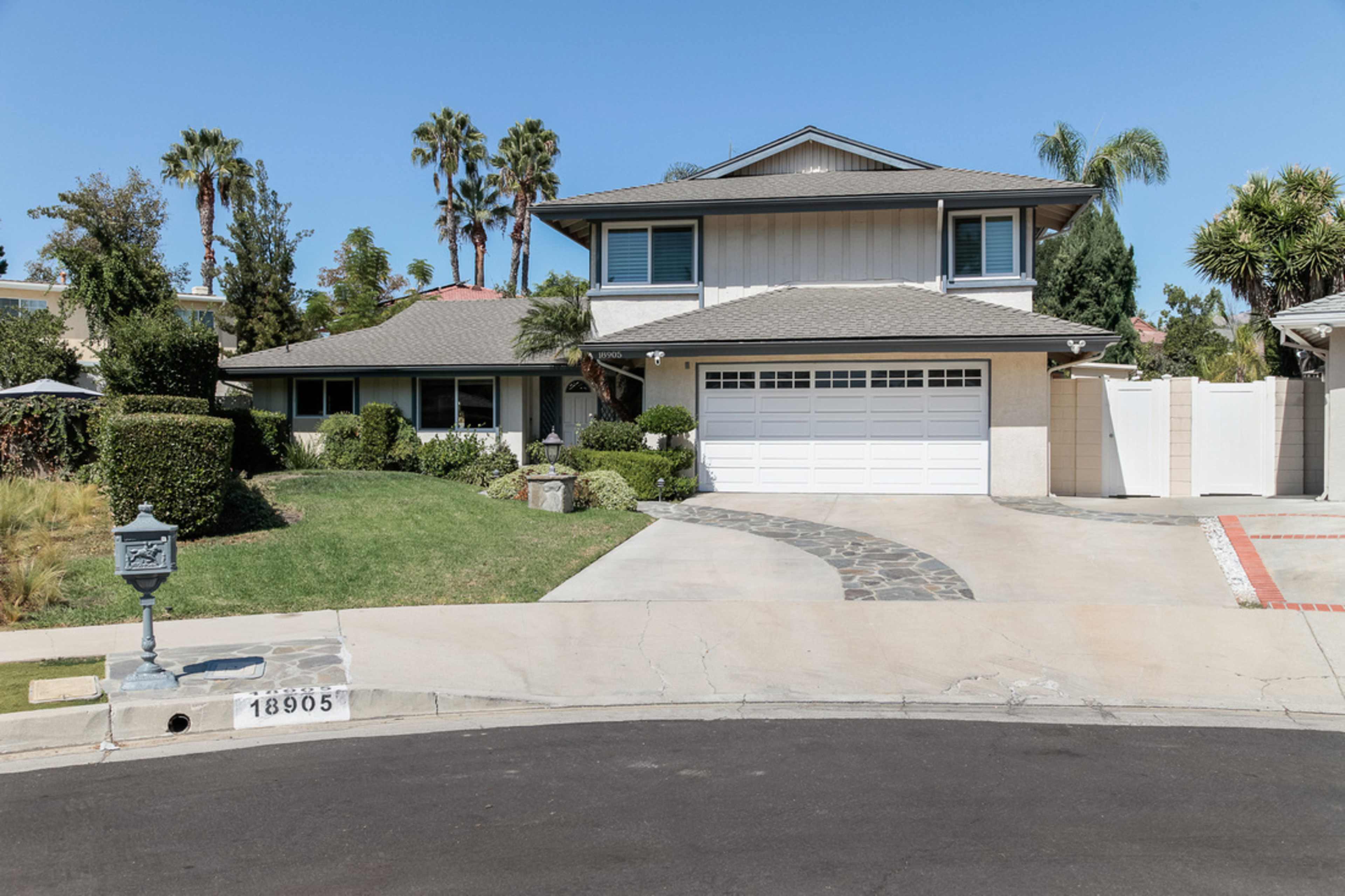 A two-story house with a gray roof and white siding sits on a landscaped corner lot with a circular driveway and palm trees.