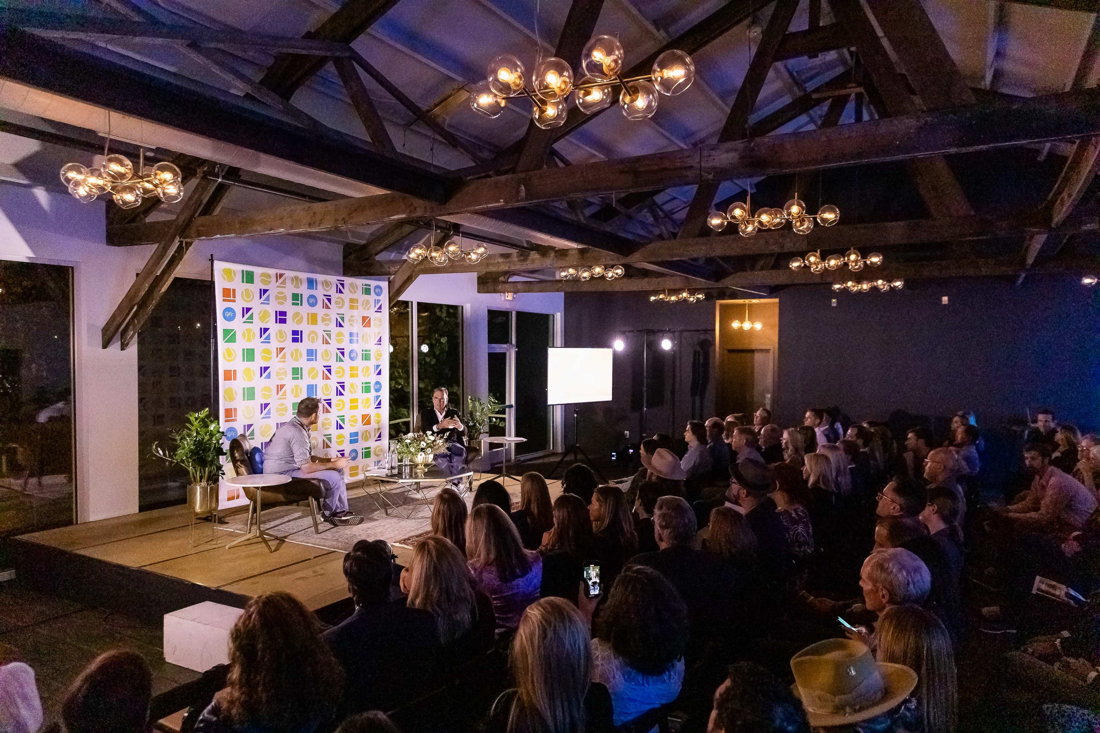 A large audience listens to two speakers on a stage in a room with exposed wooden beams and decorative pendant lights.