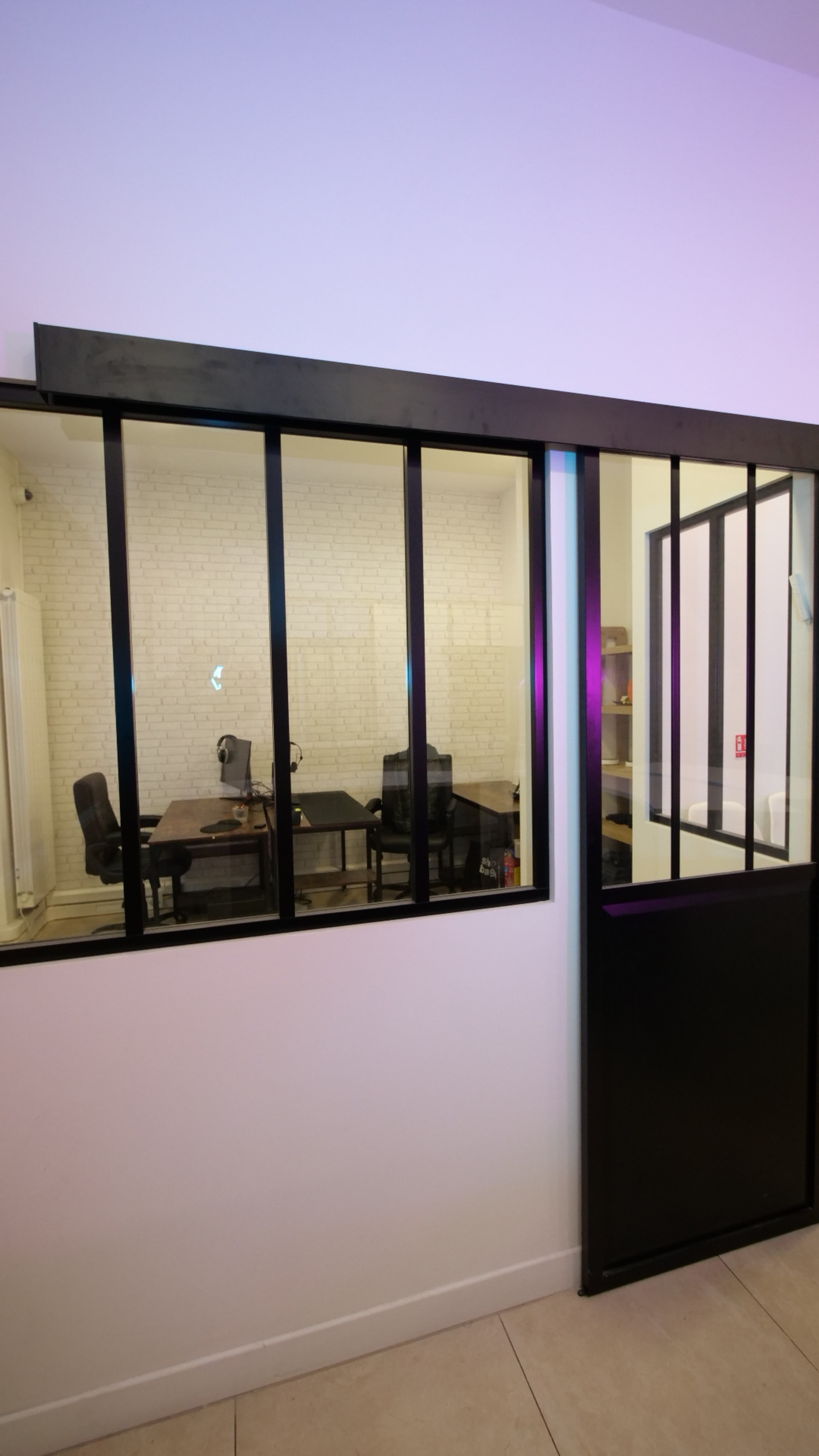 The image shows a view of a modern office space through a glass-fronted partition, featuring a minimalist design with two desks and black chairs.