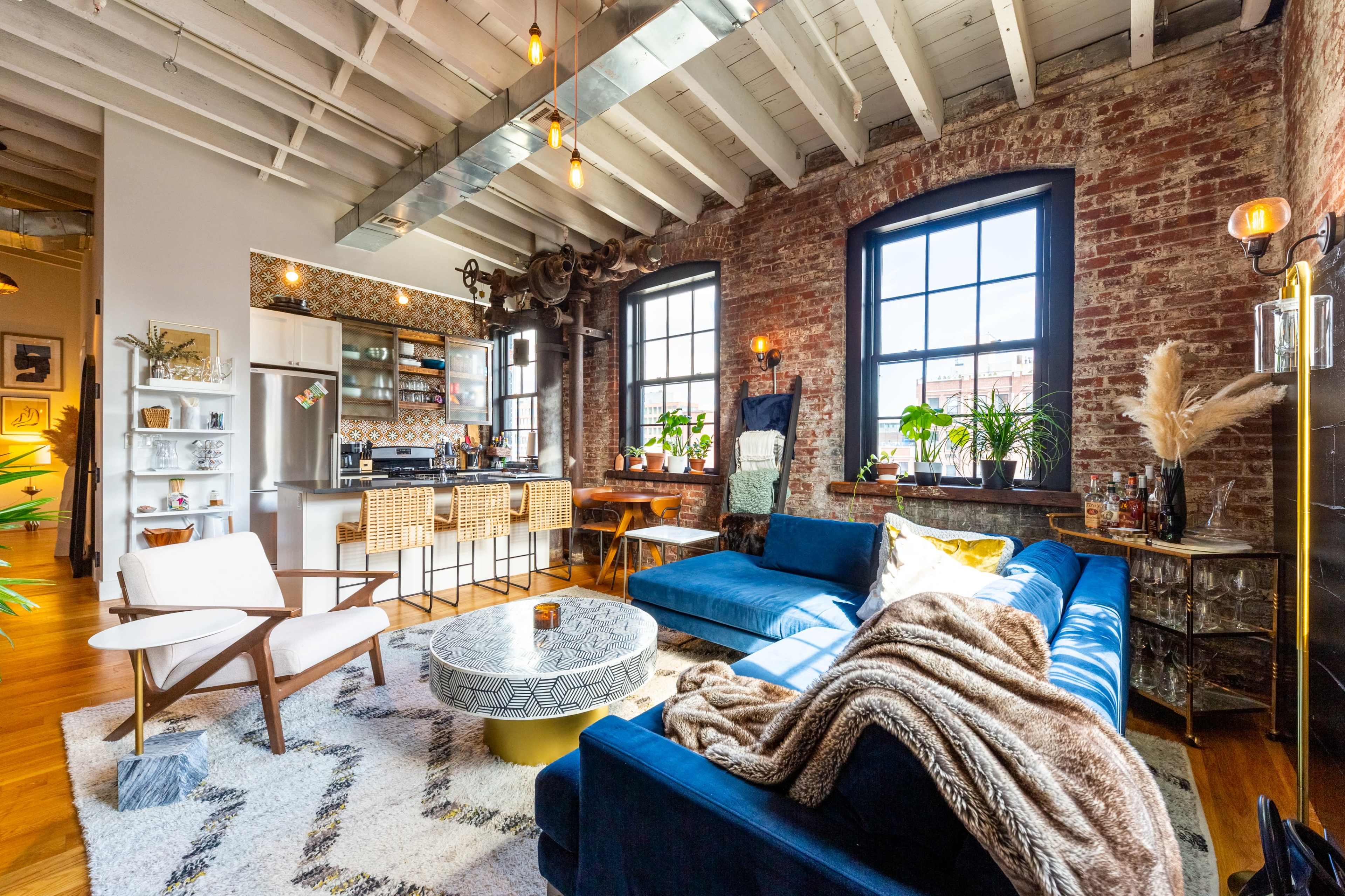 The living room features exposed brick walls, large windows, a blue sectional sofa, a patterned rug, and a kitchen area with bar stools in the background.