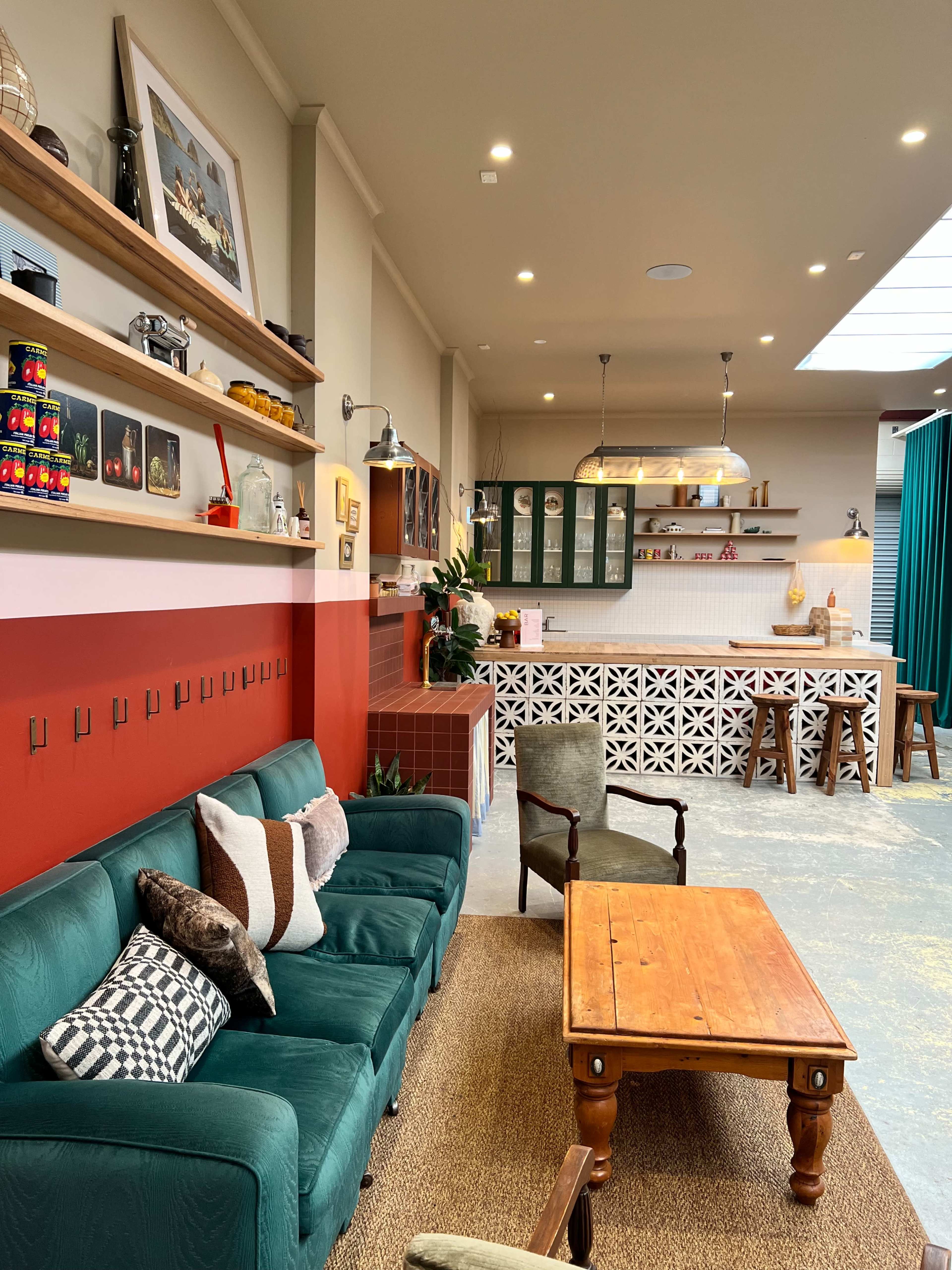The image shows a cozy interior of a cafe or lounge area featuring teal sofas, a wooden coffee table, shelves with decor items, and a well-lit kitchen space in the background.