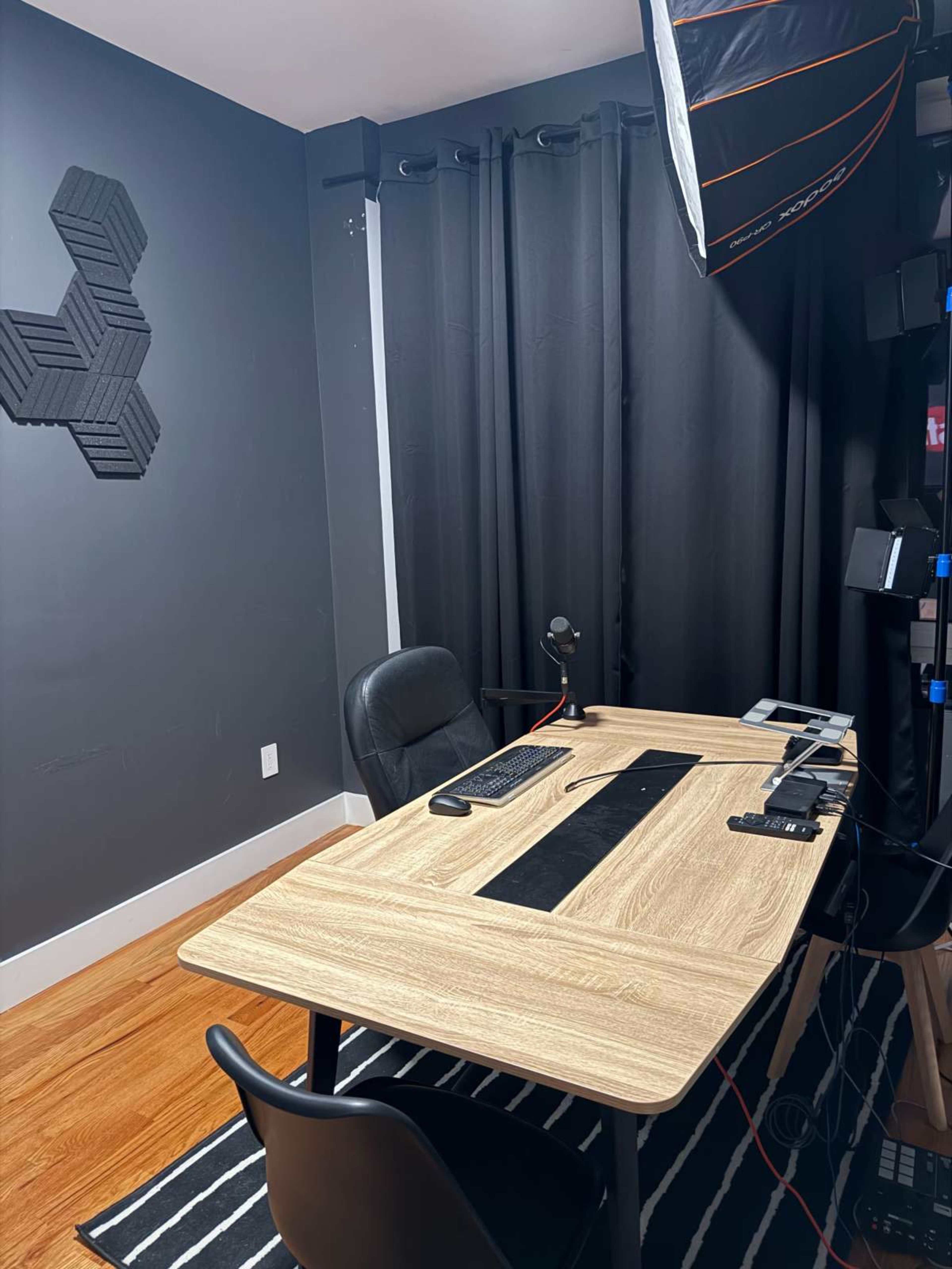 The image depicts a modern home office with a wooden desk, a black chair, and various electronic devices arranged on the desk, all set against dark walls and curtains.