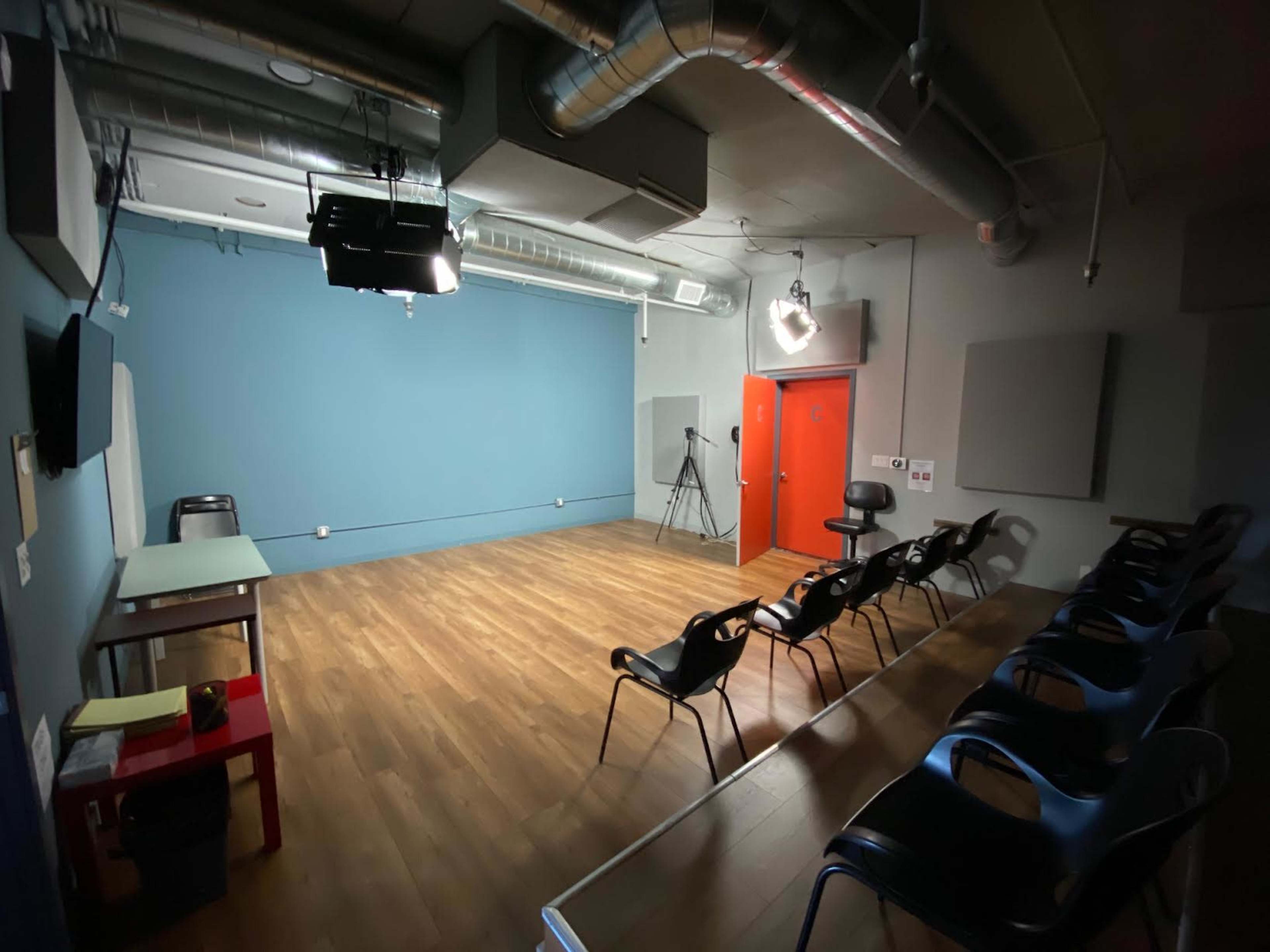An empty rehearsal studio with a blue wall, wooden flooring, a chair and table, and several black chairs arranged in a row.