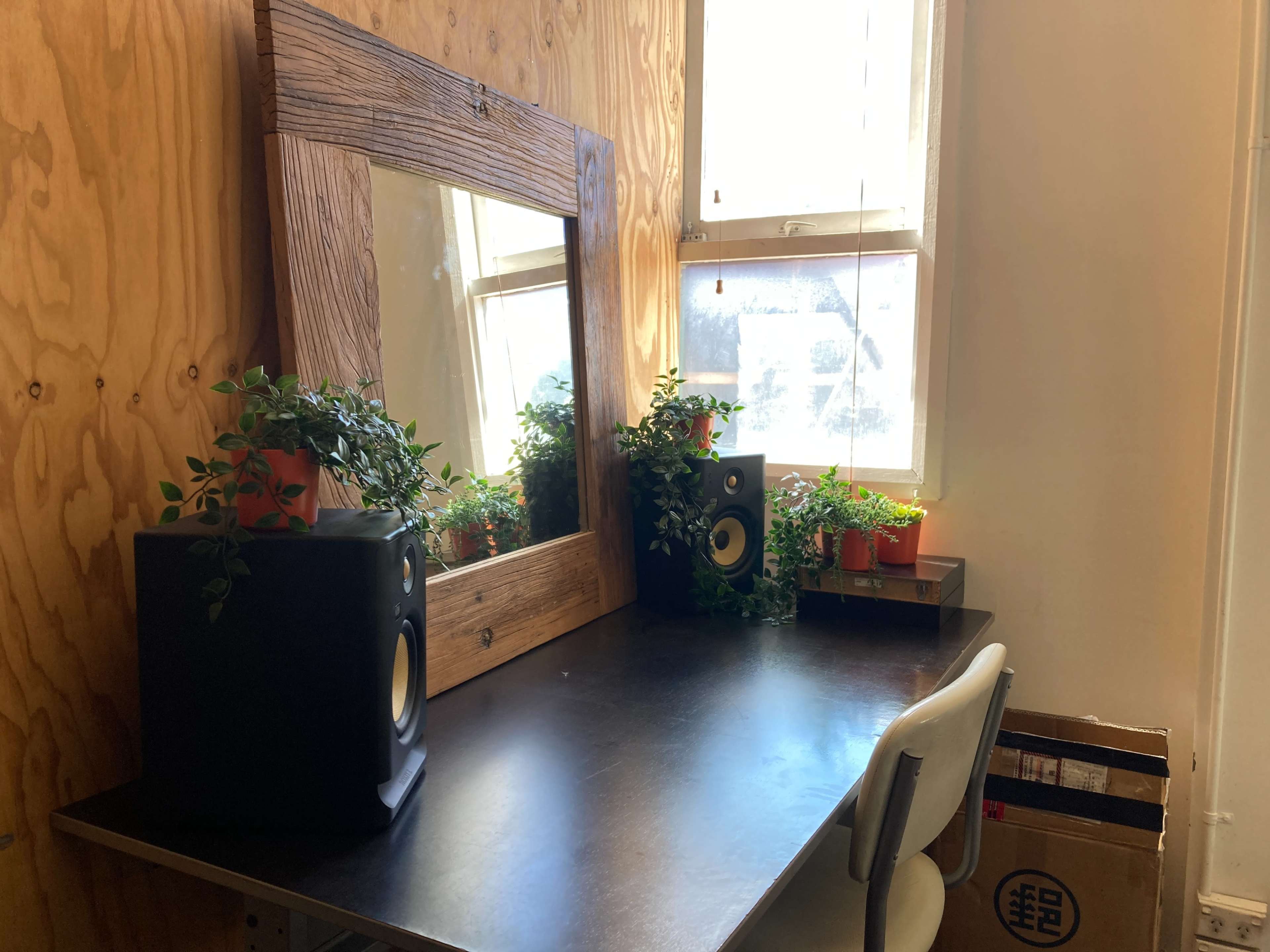 A minimalist desk with a large wooden-framed mirror, bookend speakers, and several potted plants, set against a wooden wall.