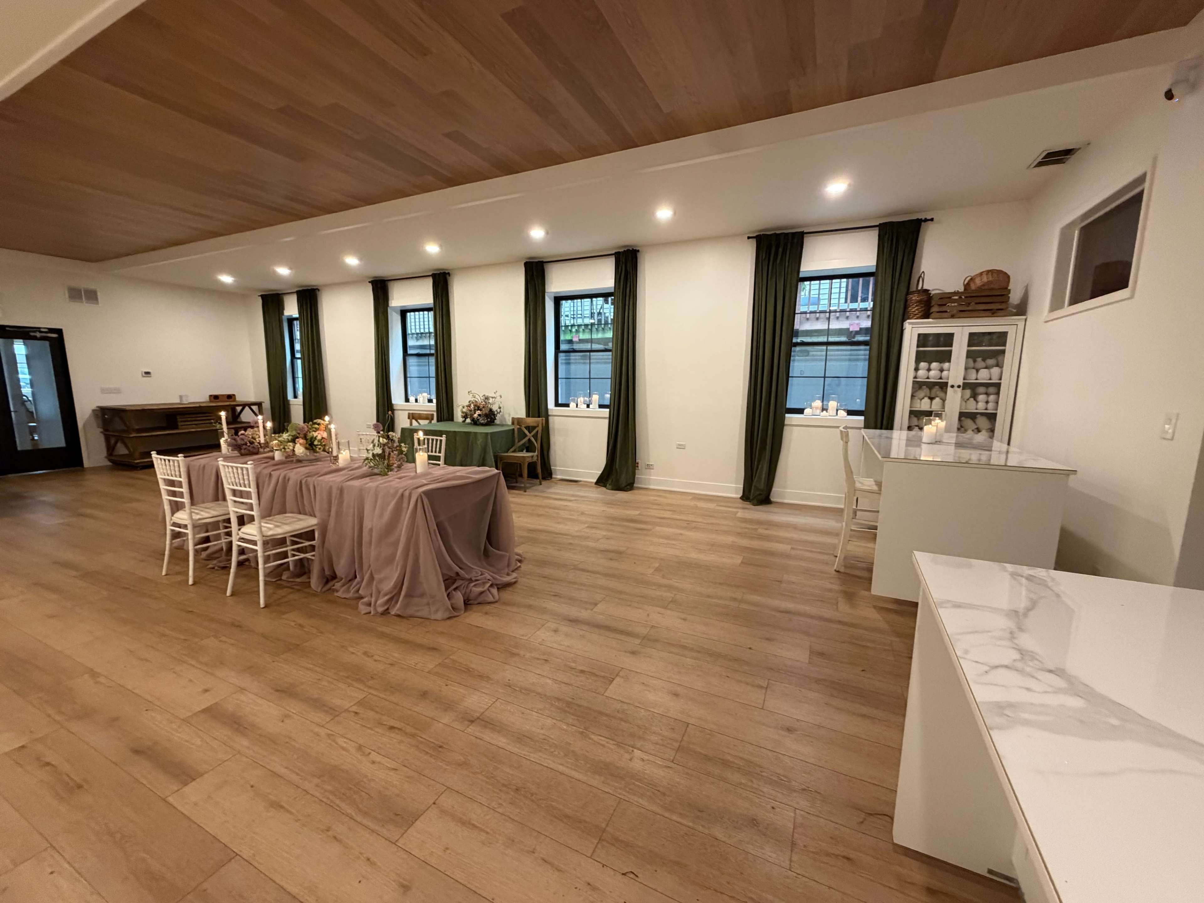 The image shows a spacious room with wooden flooring, featuring a long table set with a tablecloth and dining decor alongside additional seating areas and a white furniture piece against the wall.