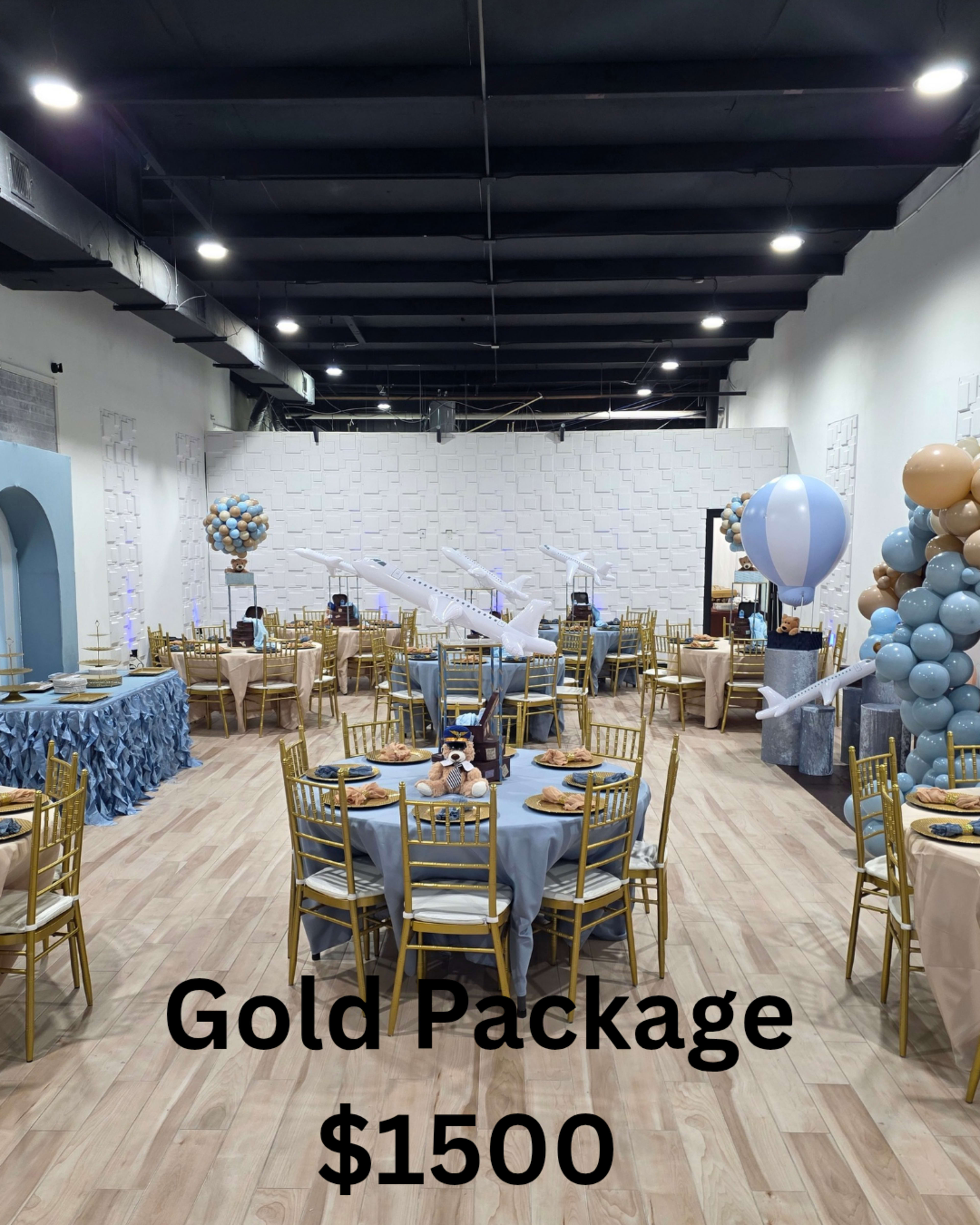 The image shows a well-decorated event space set up for a gathering, featuring round tables with blue tablecloths, gold chairs, and decorative balloons.