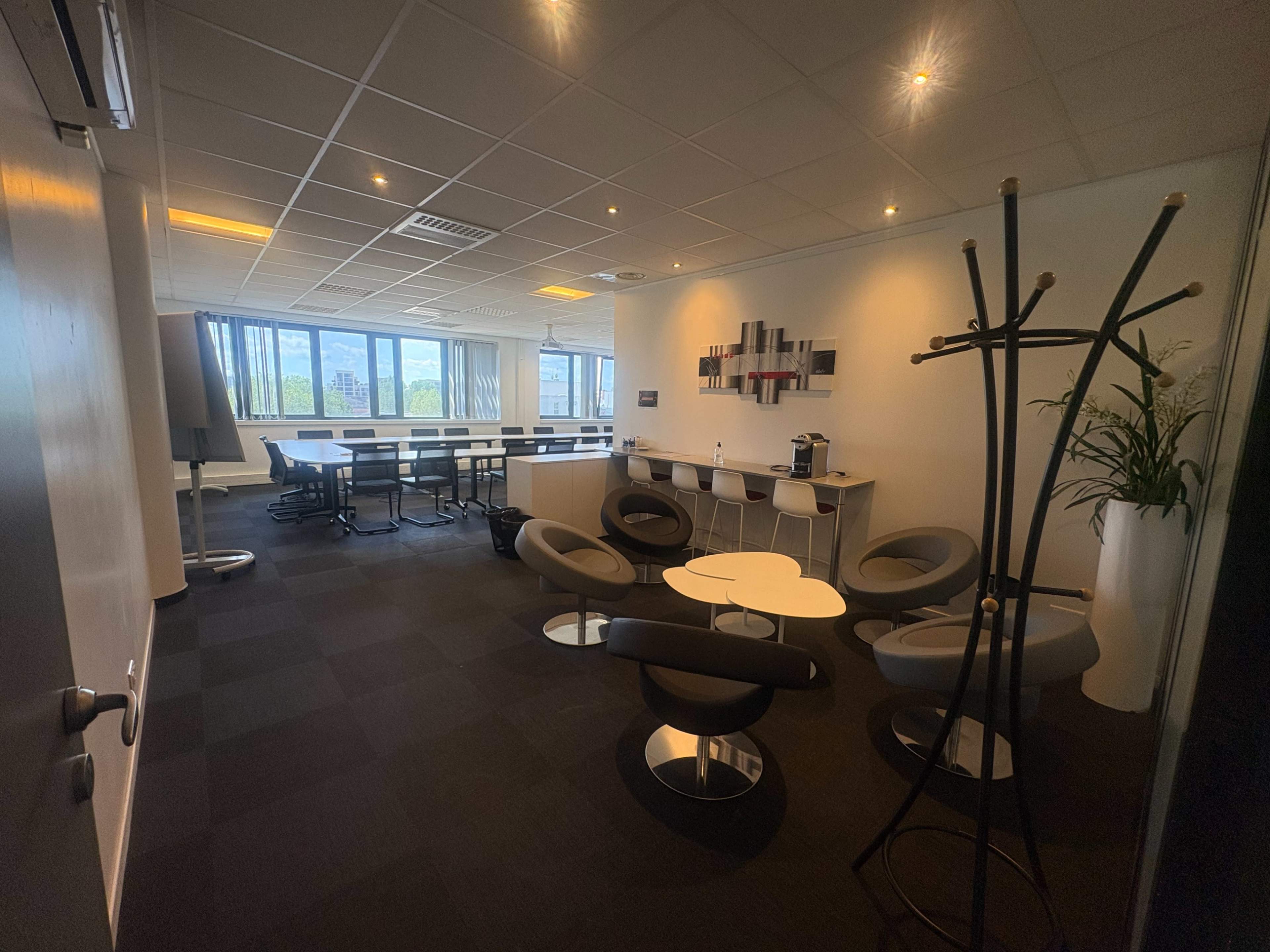 The image shows a modern office meeting room with seating areas and large windows providing natural light.