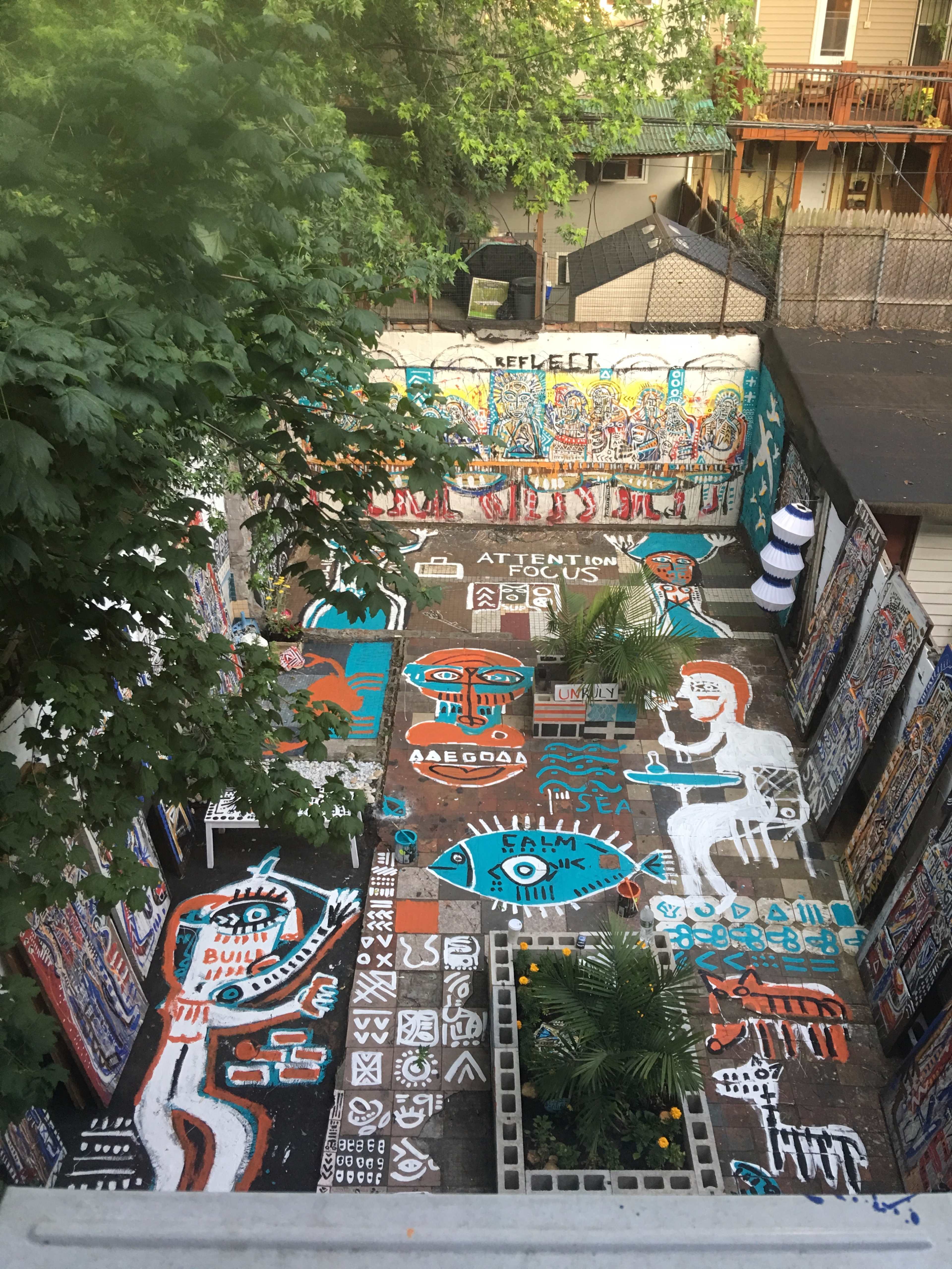 The image shows a vibrant outdoor space covered with colorful street art and murals, surrounded by greenery and buildings.