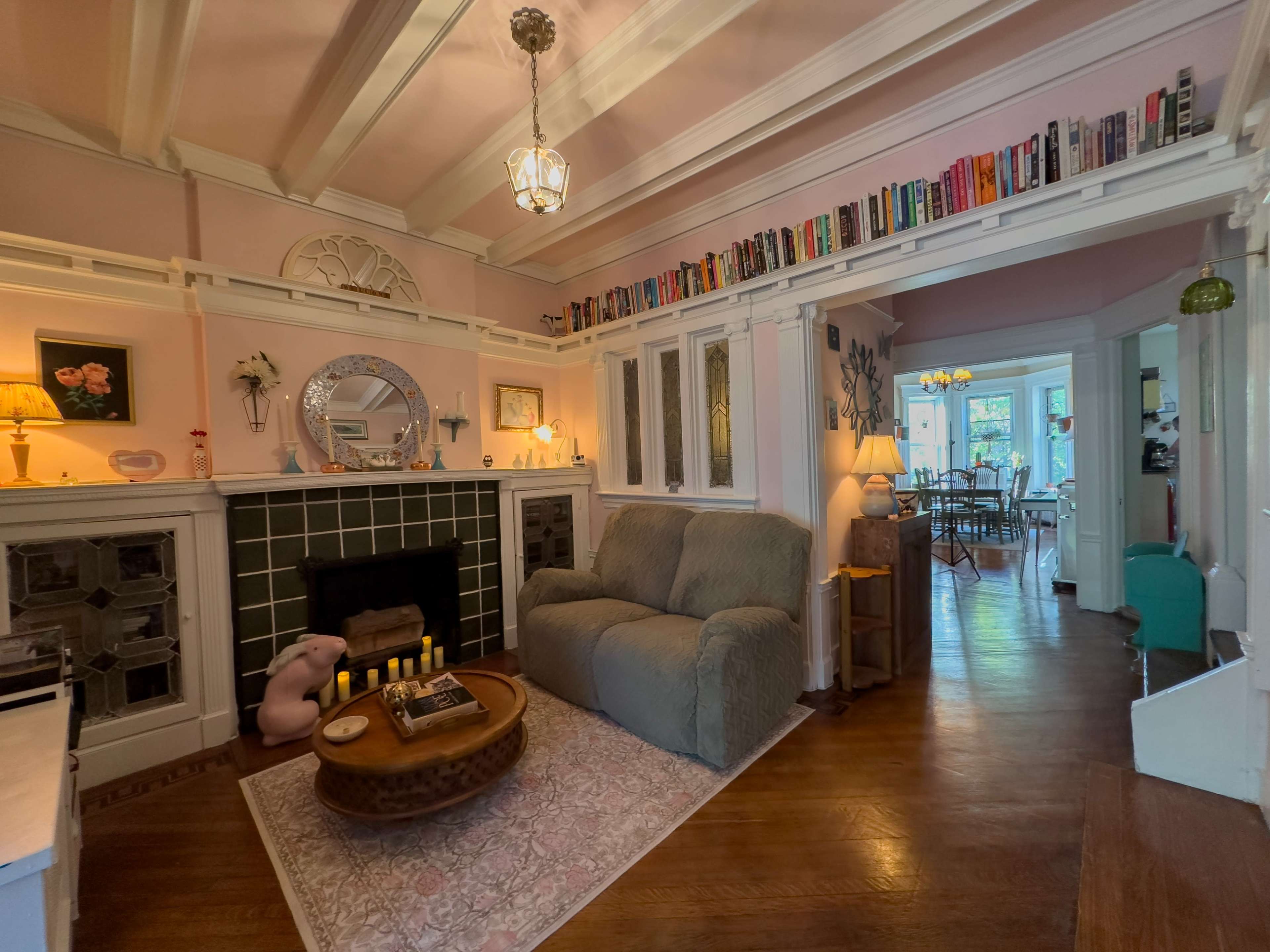 The image shows a cozy living room with a light pink color scheme, featuring a fireplace, a comfortable sofa, and a collection of books on an upper shelf.