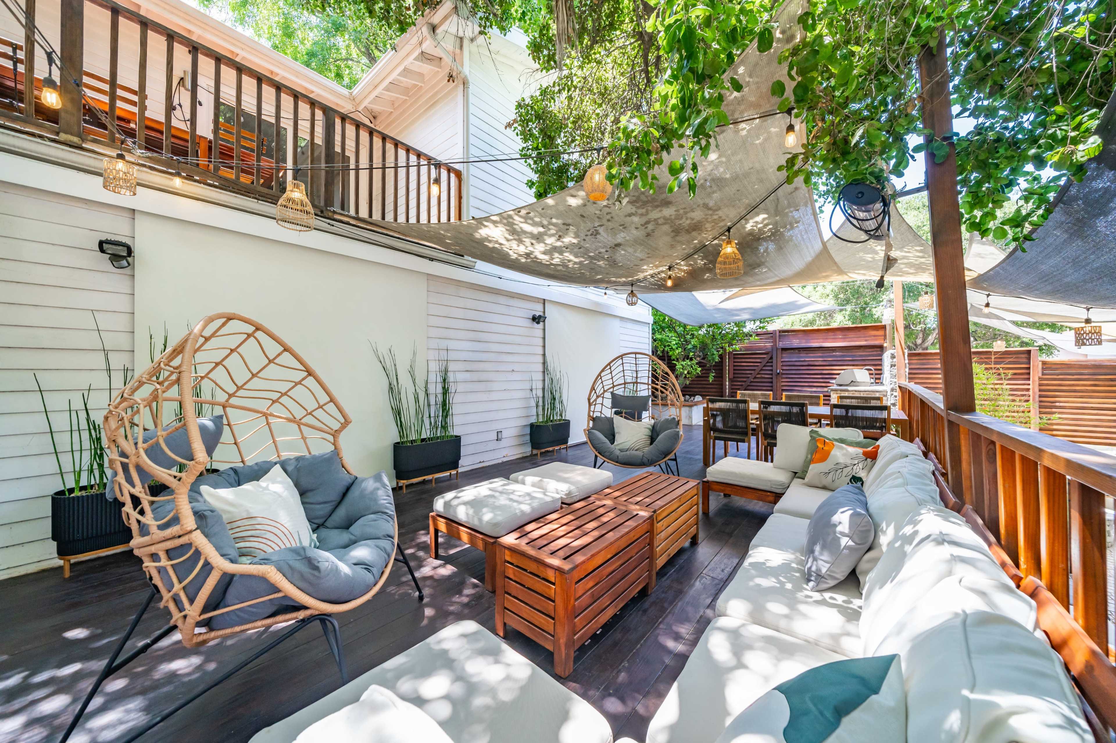 The image shows a landscaped outdoor seating area with woven chairs, a wooden coffee table, and shade provided by overhead fabric and greenery.