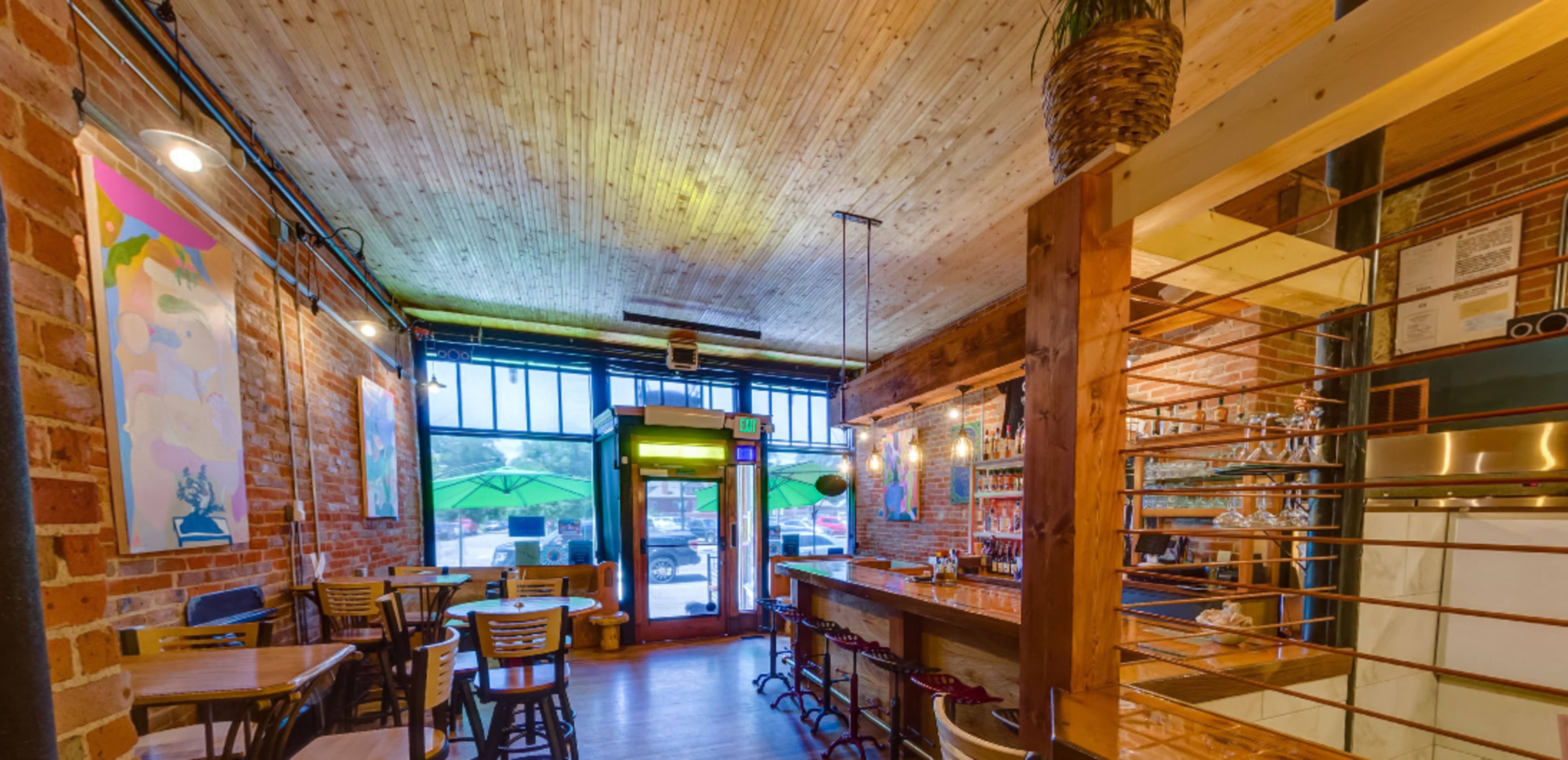 A modern café with exposed brick walls, wooden beams, and a bar area, featuring tables and chairs arranged along the side.