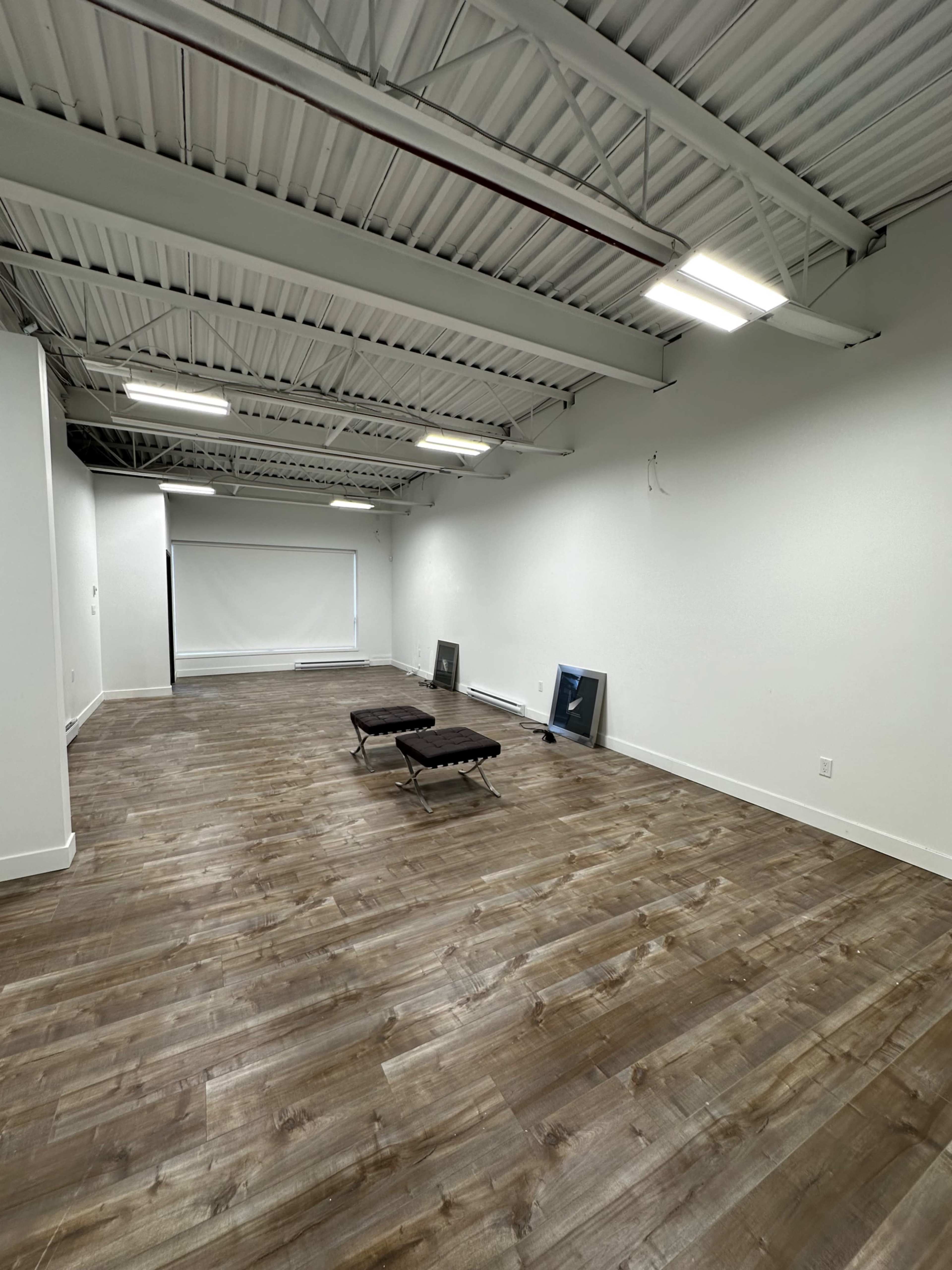 The image shows an empty room with wooden flooring, two folding chairs, and white walls, featuring a projector screen at one end.