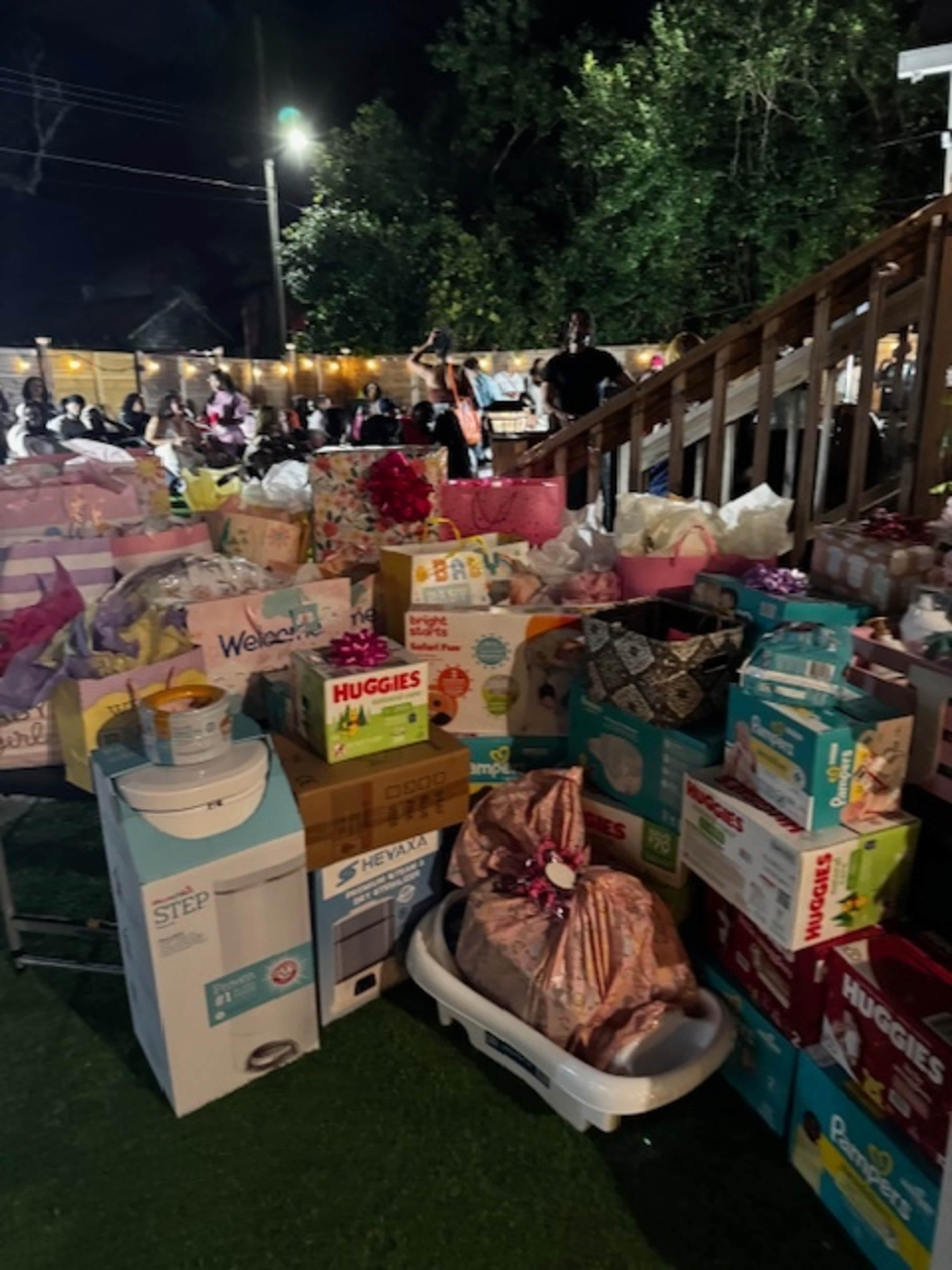 The scene shows a large gathering at night with many tables filled with colorful gift boxes and baby supplies, surrounded by people in the background.