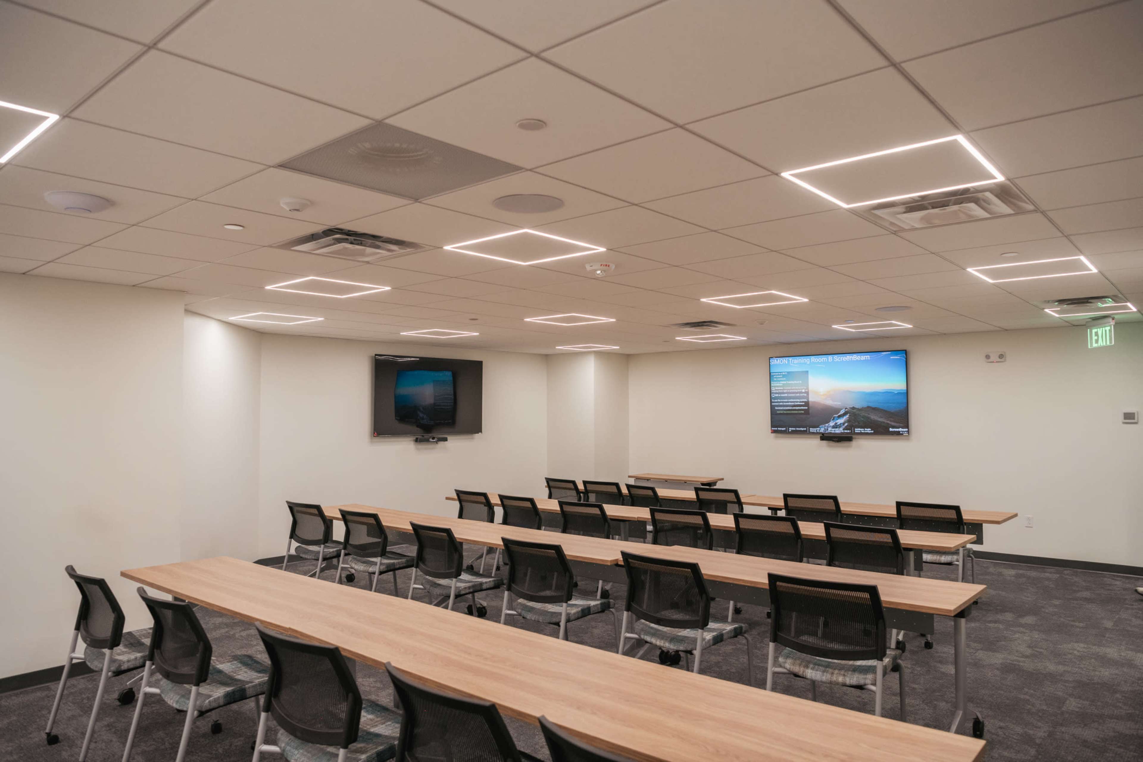 The image shows a modern conference room with rows of tables and chairs, equipped with two screens mounted on the walls.