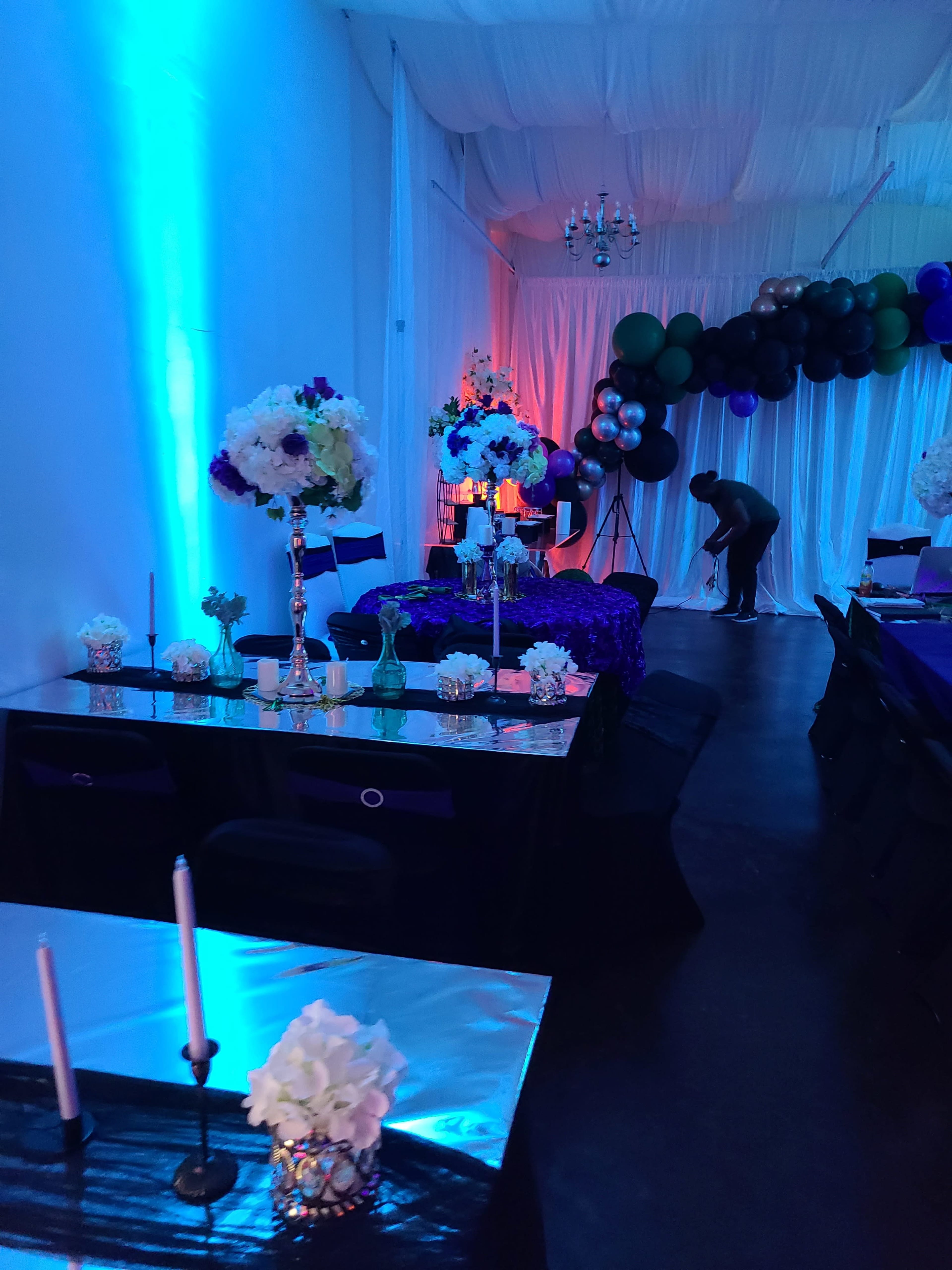 A decorated indoor event space features tables with floral centerpieces, candles, and colorful lighting along with a backdrop of balloons.