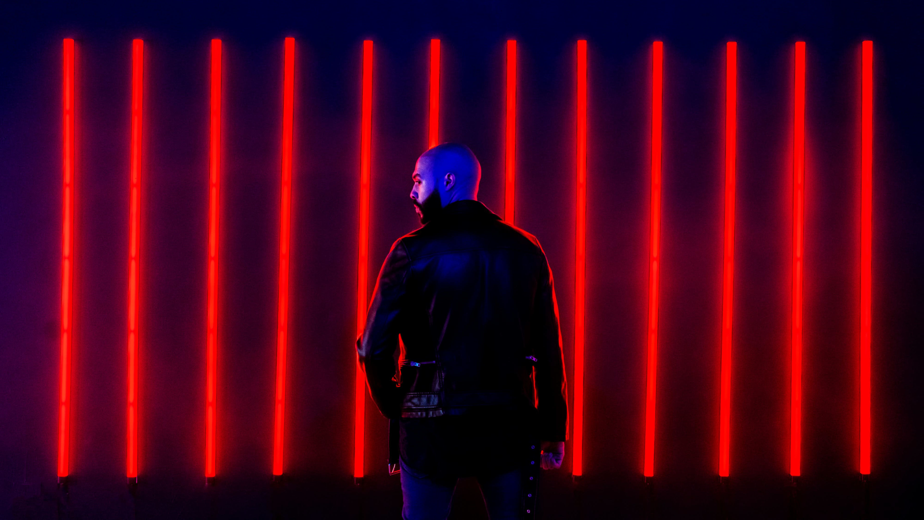 A person in a black leather jacket stands with their back to a wall of vertical red neon lights.