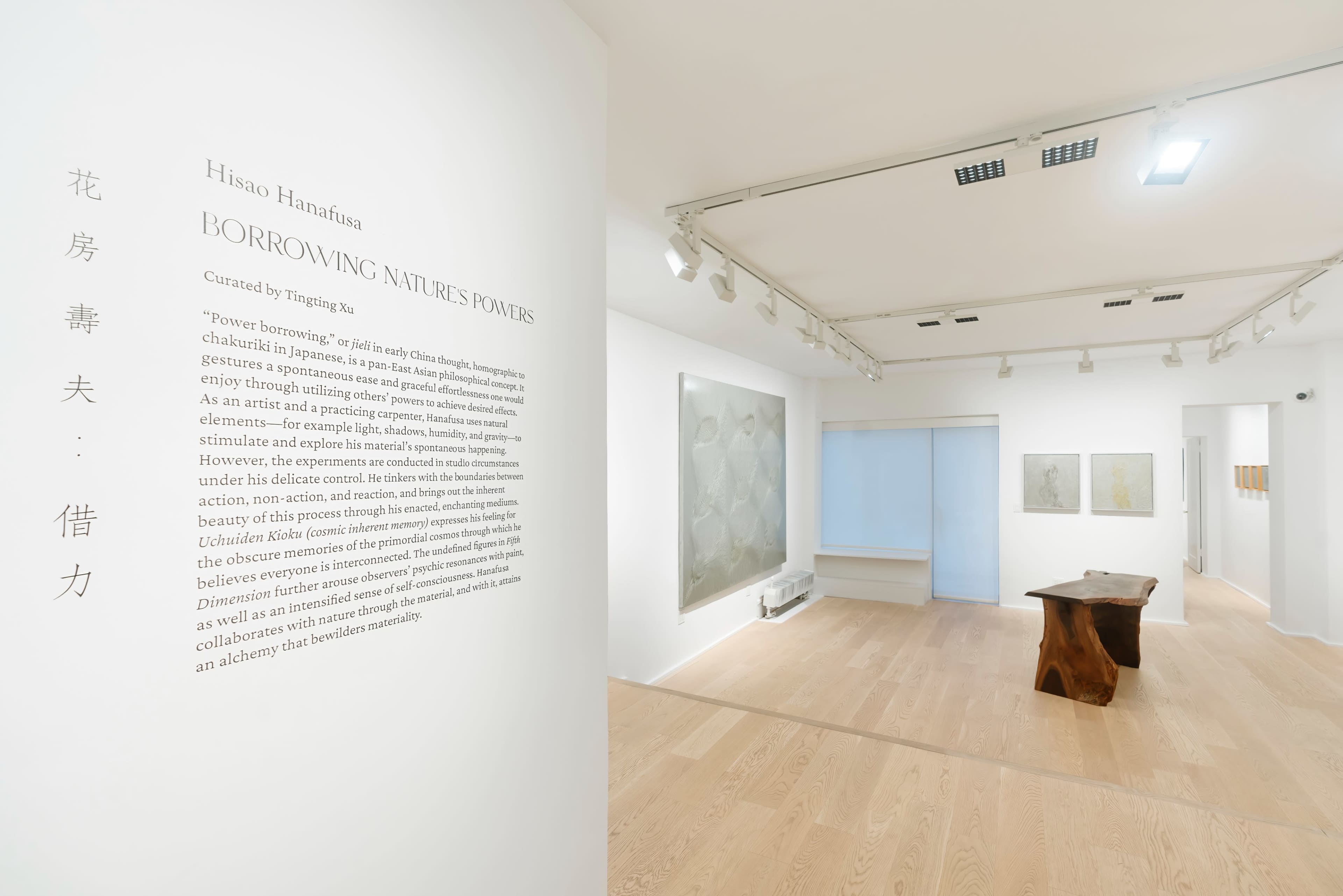 A minimalist art gallery featuring a large text panel on the left wall and various artworks displayed on the right, along with a wooden table in the foreground.