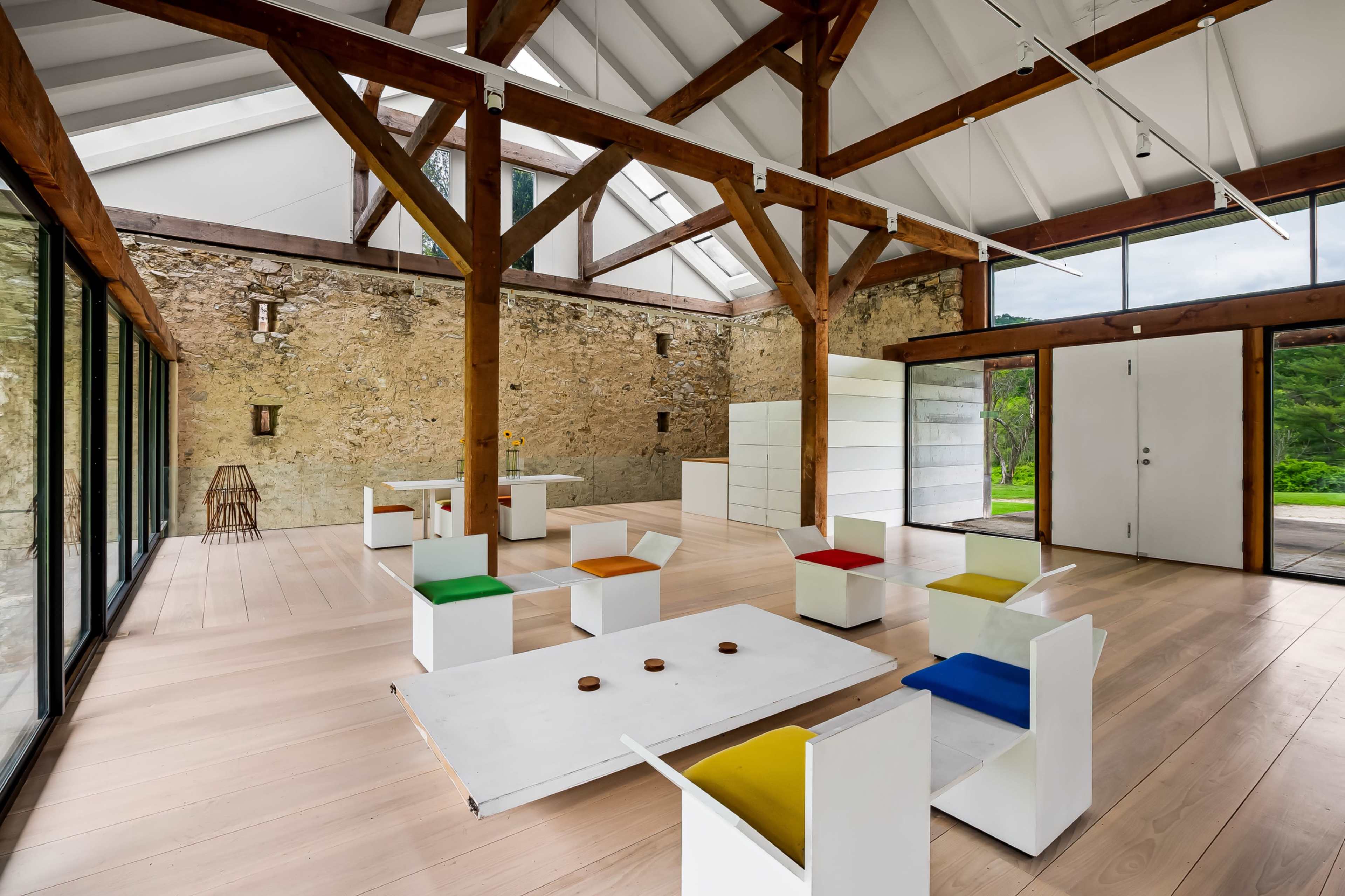 The image shows a modern interior space featuring wooden beams, a stone wall, and colorful seating arrangements around a central table.