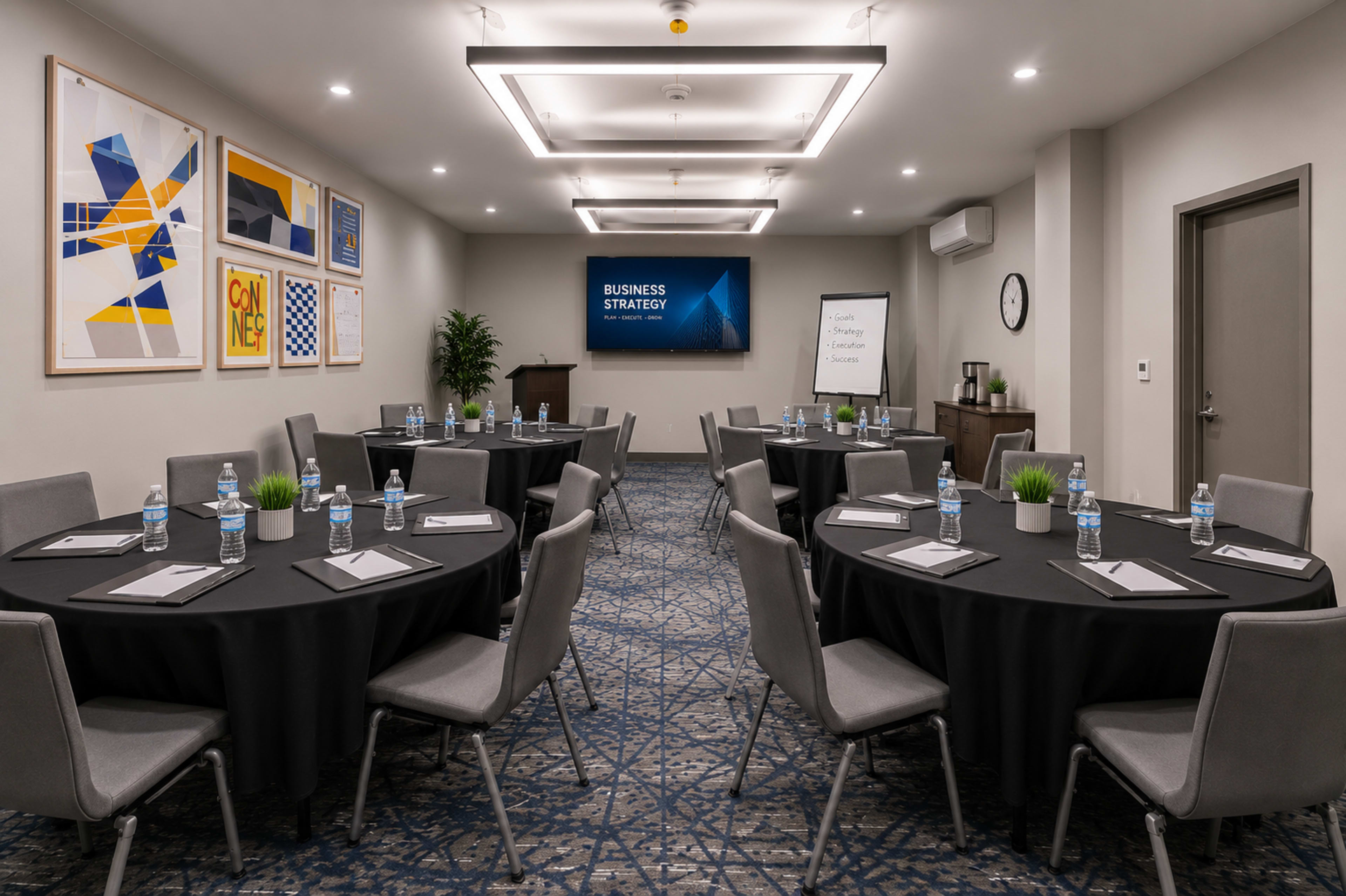 The image shows a conference room set up with several round tables covered in black tablecloths, each with water bottles and notepads, and a large screen displaying "Business Strategy" at the front.