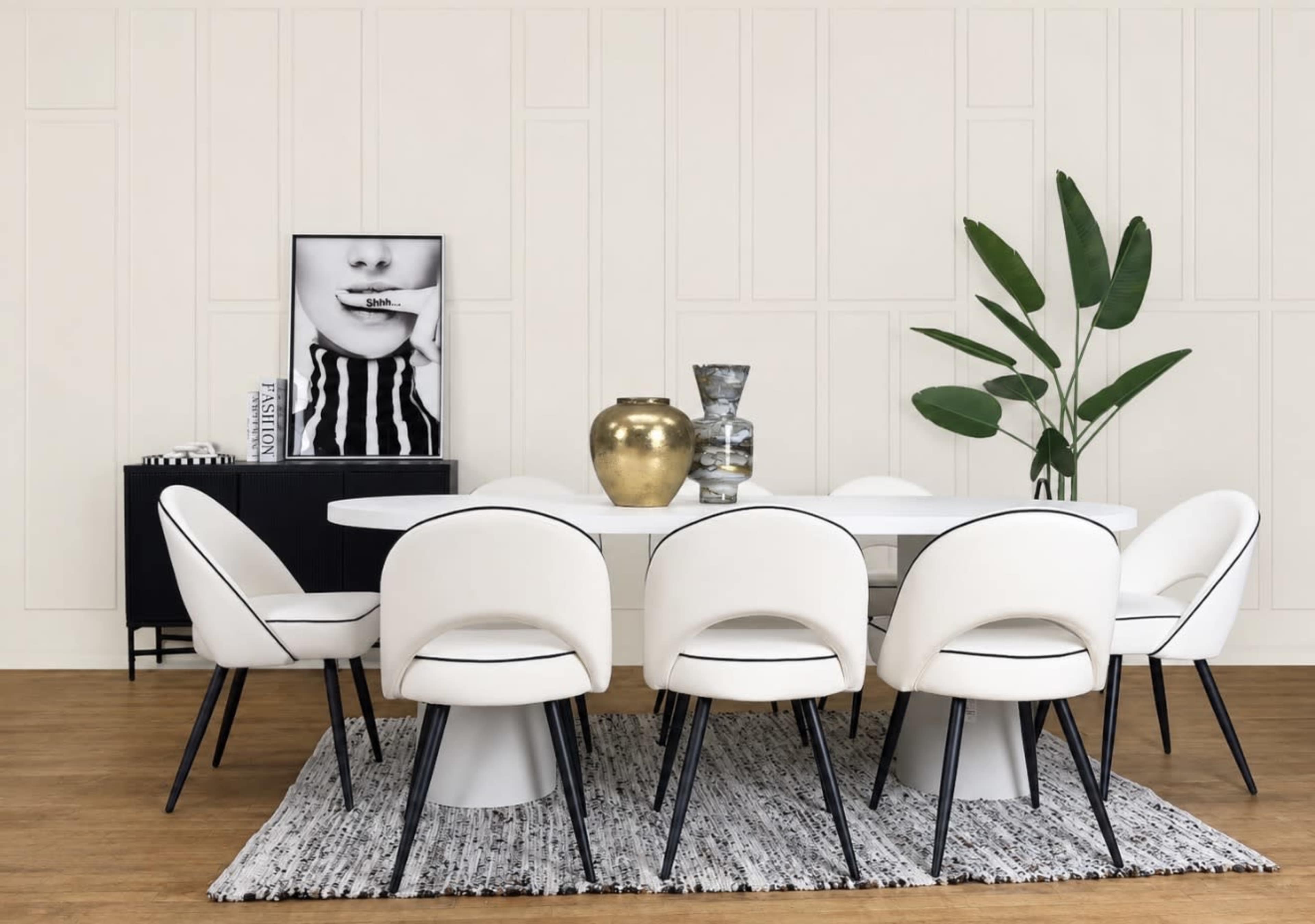 The image shows a modern dining room setup featuring a round table surrounded by eight white upholstered chairs, with decorative items on the table and a black cabinet against the wall.