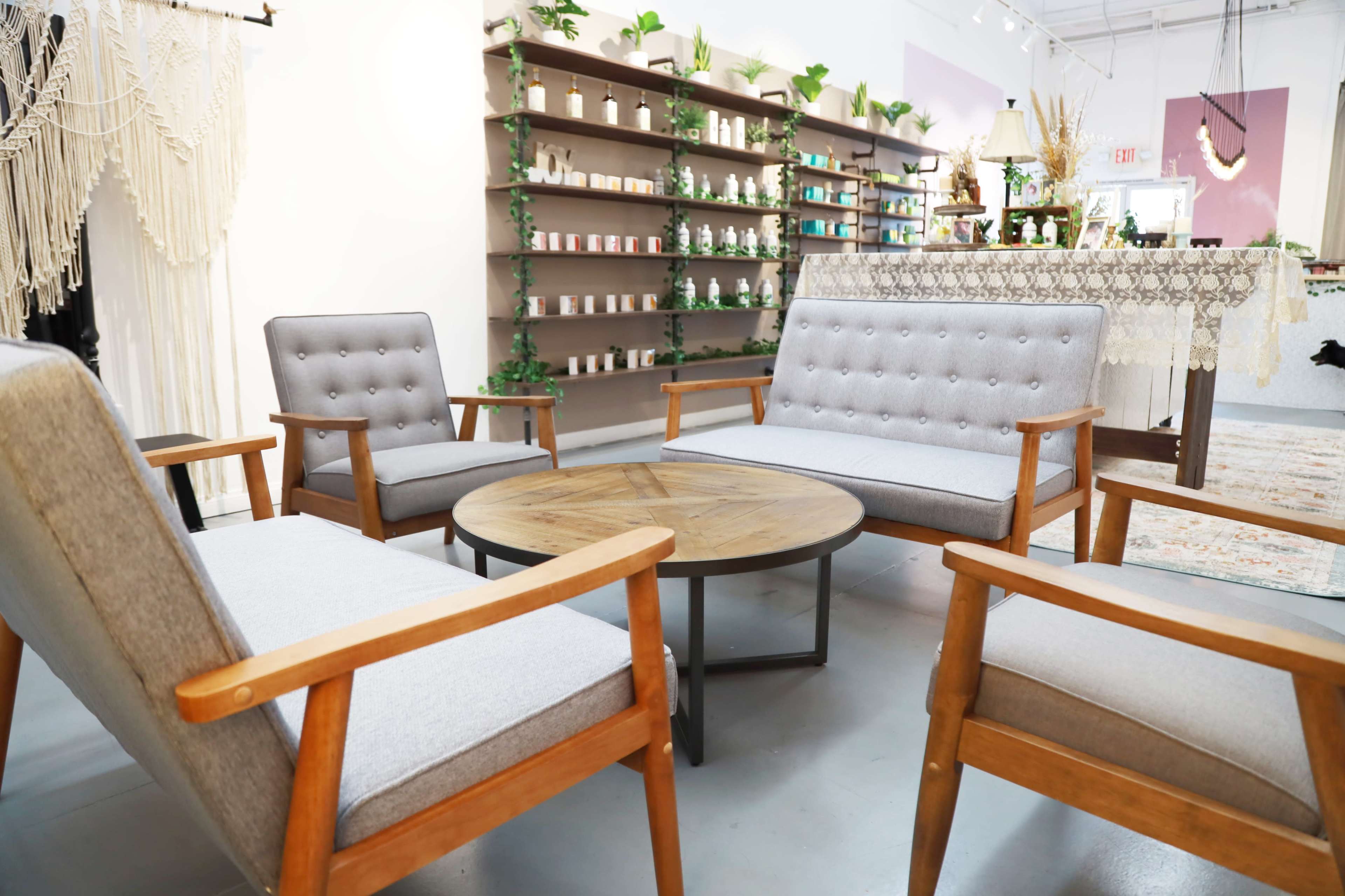 A seating area features four wooden chairs with gray upholstery arranged around a circular wooden table, set against a backdrop of shelves displaying decorative items and plants.