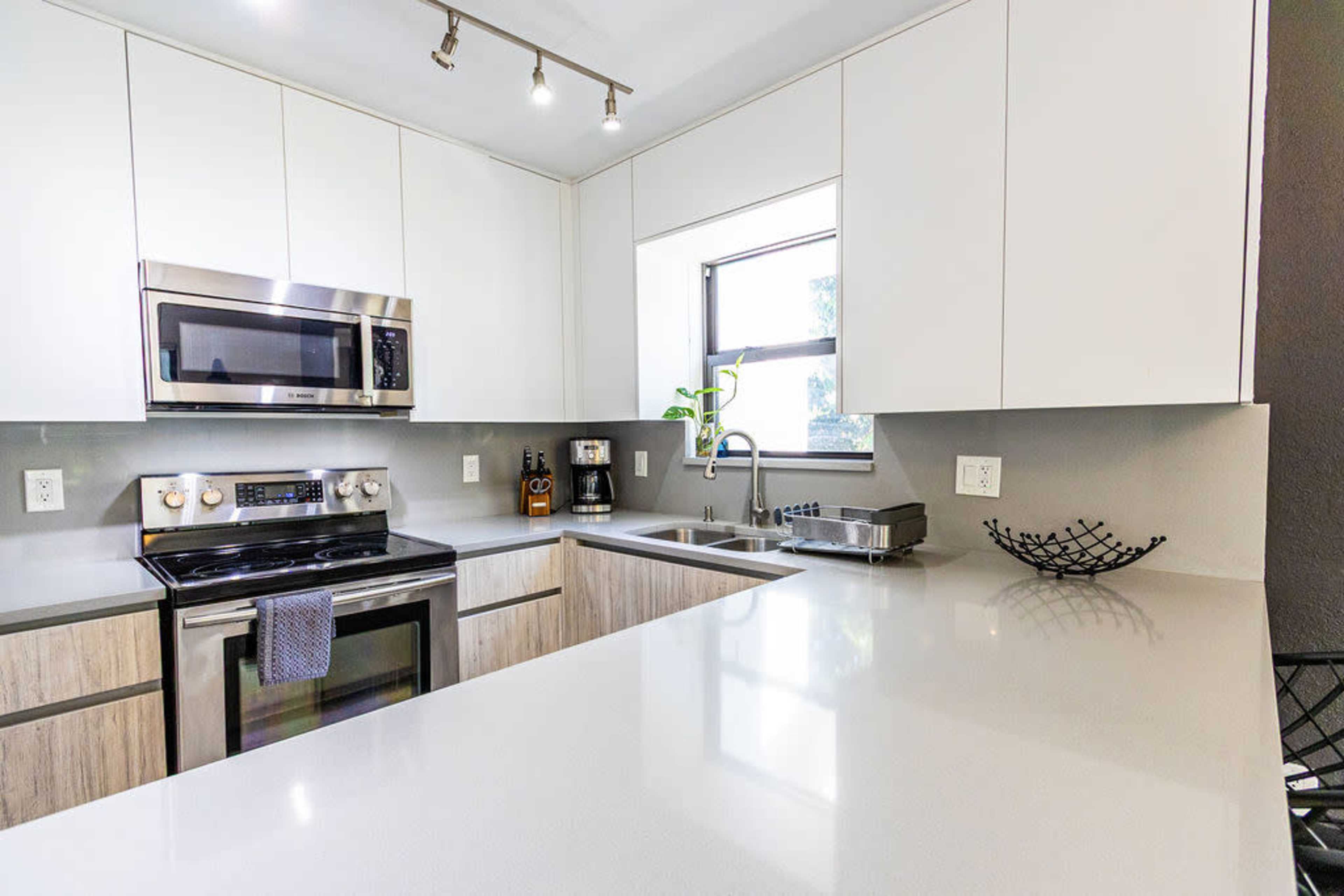 The image shows a modern kitchen with stainless steel appliances, a large countertop, and a window allowing natural light.
