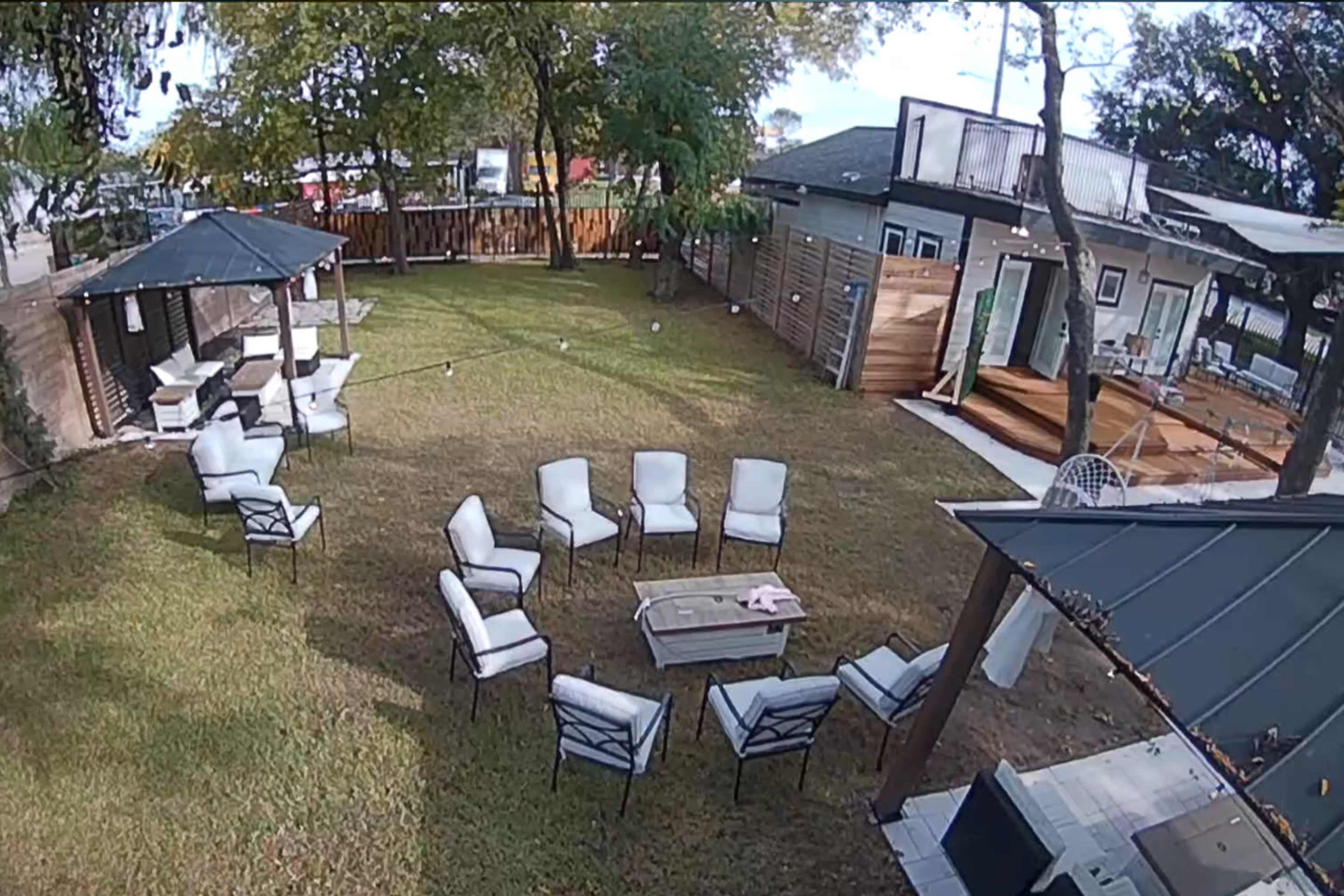 A landscaped backyard with several seating arrangements, including white chairs arranged in a circle around a table, surrounded by trees and buildings.
