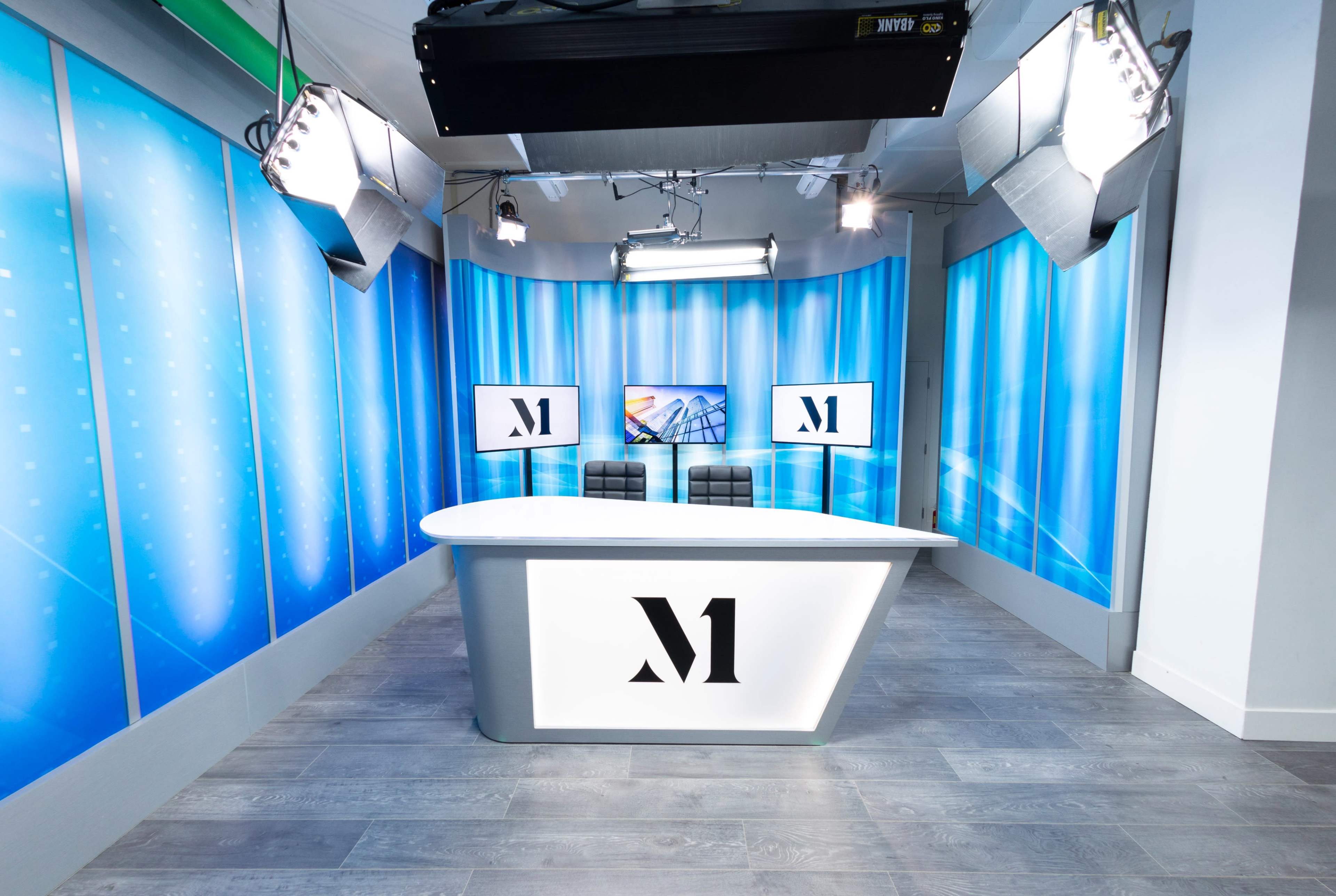 A modern television studio featuring a sleek, white desk with an "M" logo, flanked by two monitors and illuminated by overhead lights against a blue backdrop.