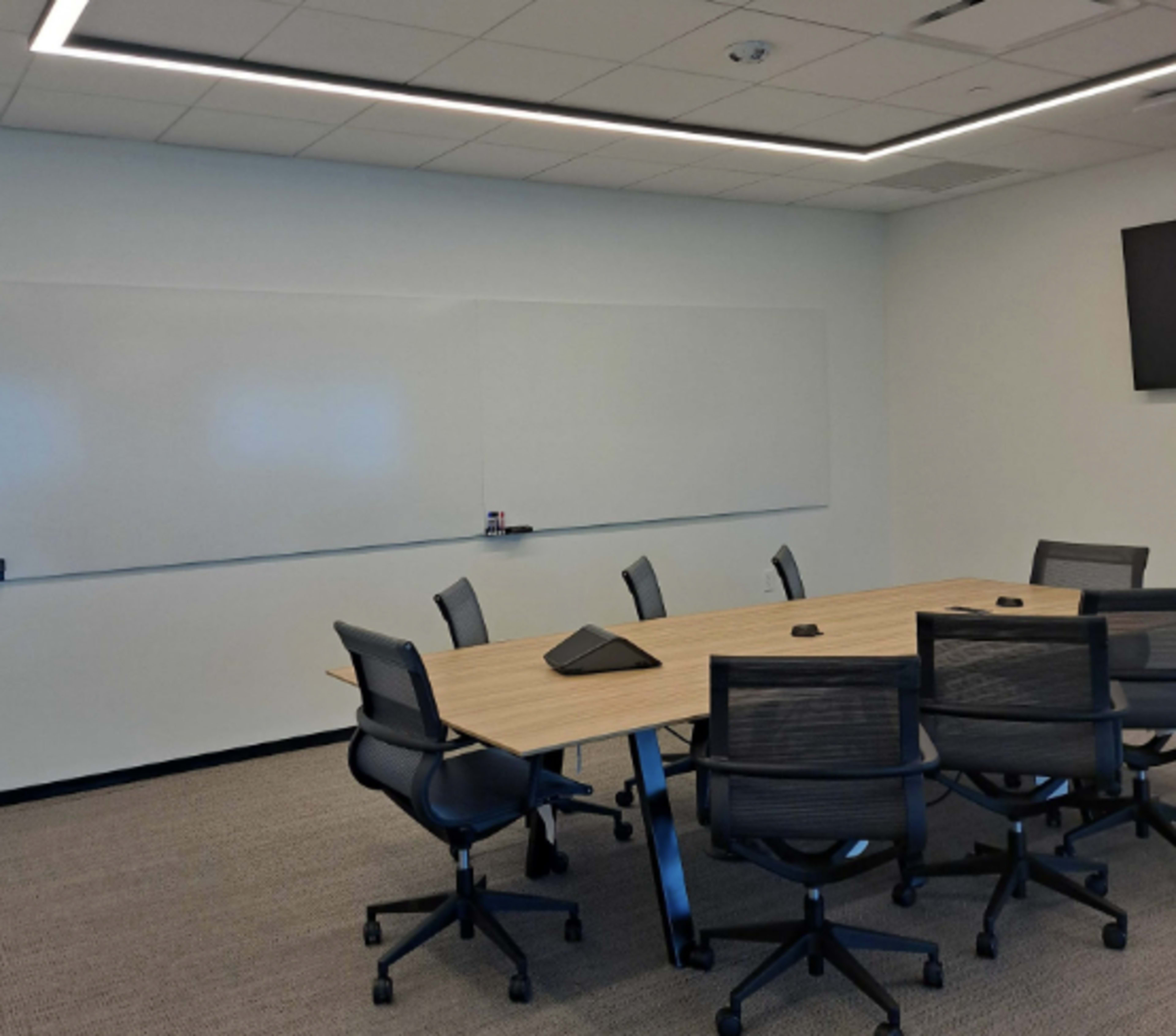 A modern conference room with a rectangular wooden table surrounded by black mesh chairs, whiteboards on the walls, and a mounted television.