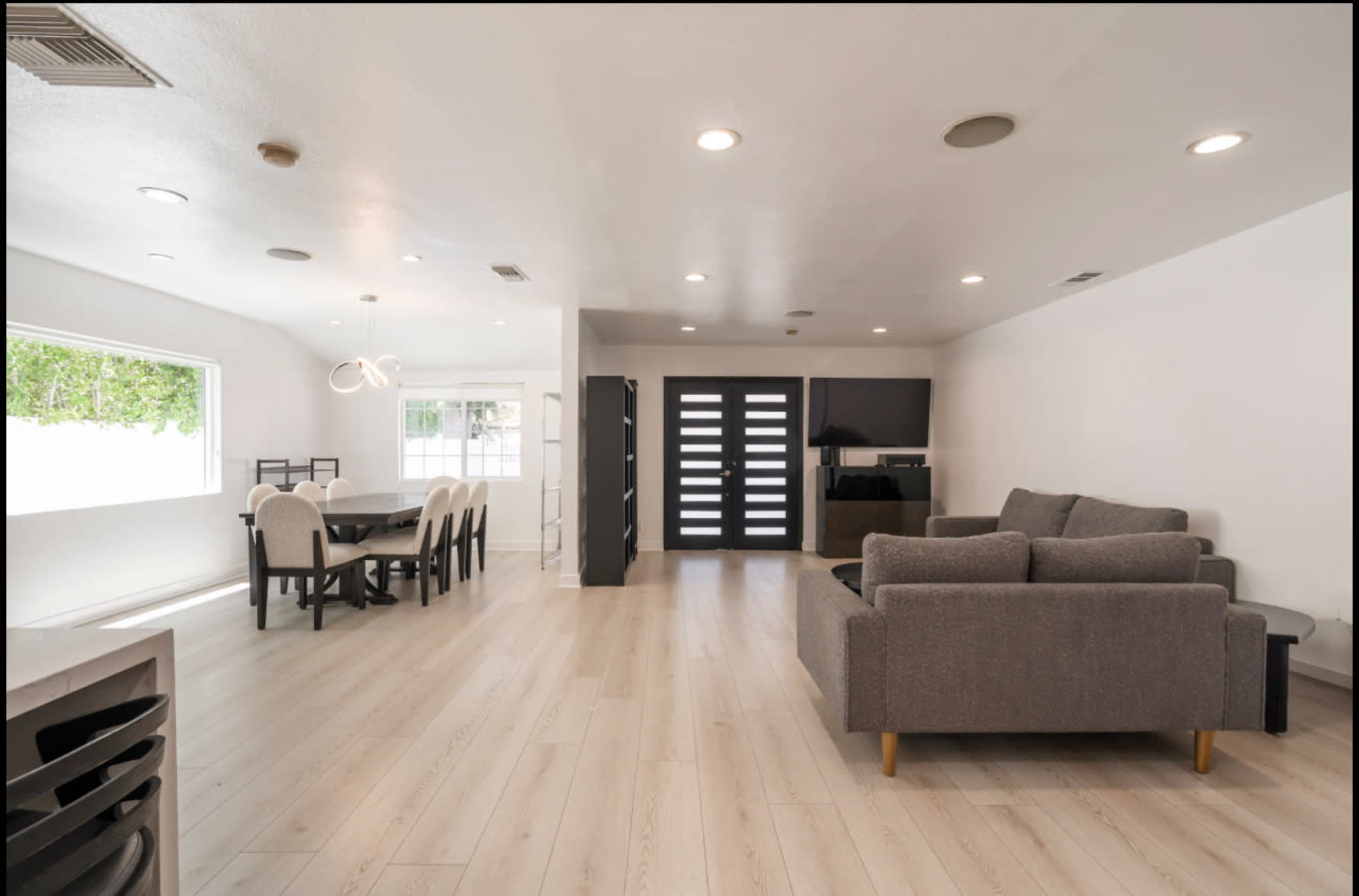 The image shows a modern interior living space featuring a dining area with a black table and six chairs, a gray sofa, and large windows allowing natural light.