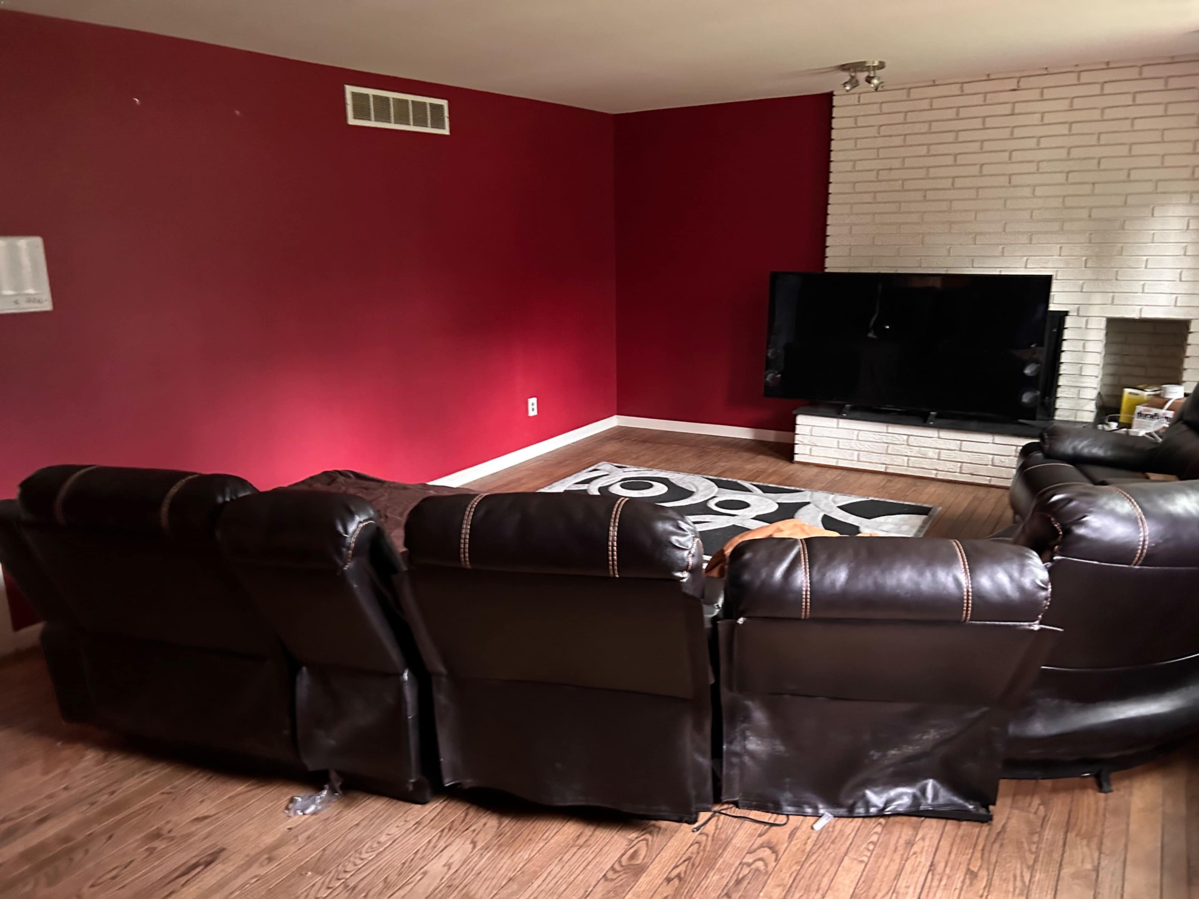 A brown leather sectional couch faces a large television mounted on a white brick wall in a room with red walls.