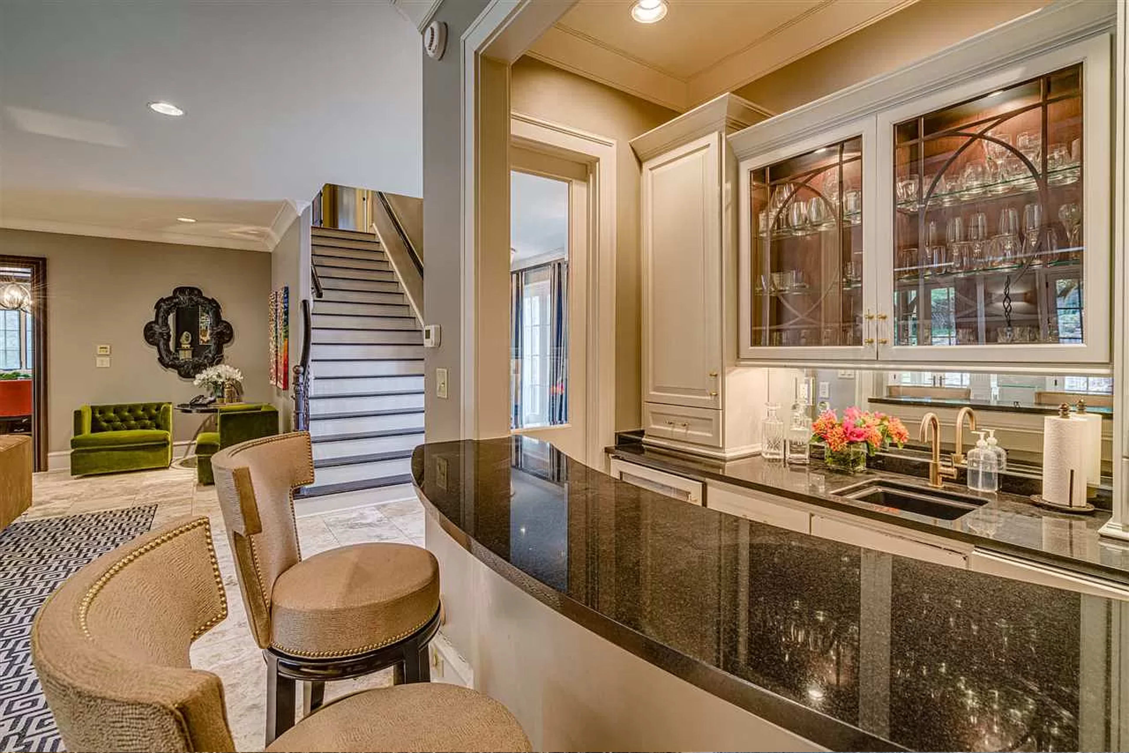 A bar area with a black countertop, light-colored cabinetry, and glass shelves, adjacent to a staircase and a living area with green seating.