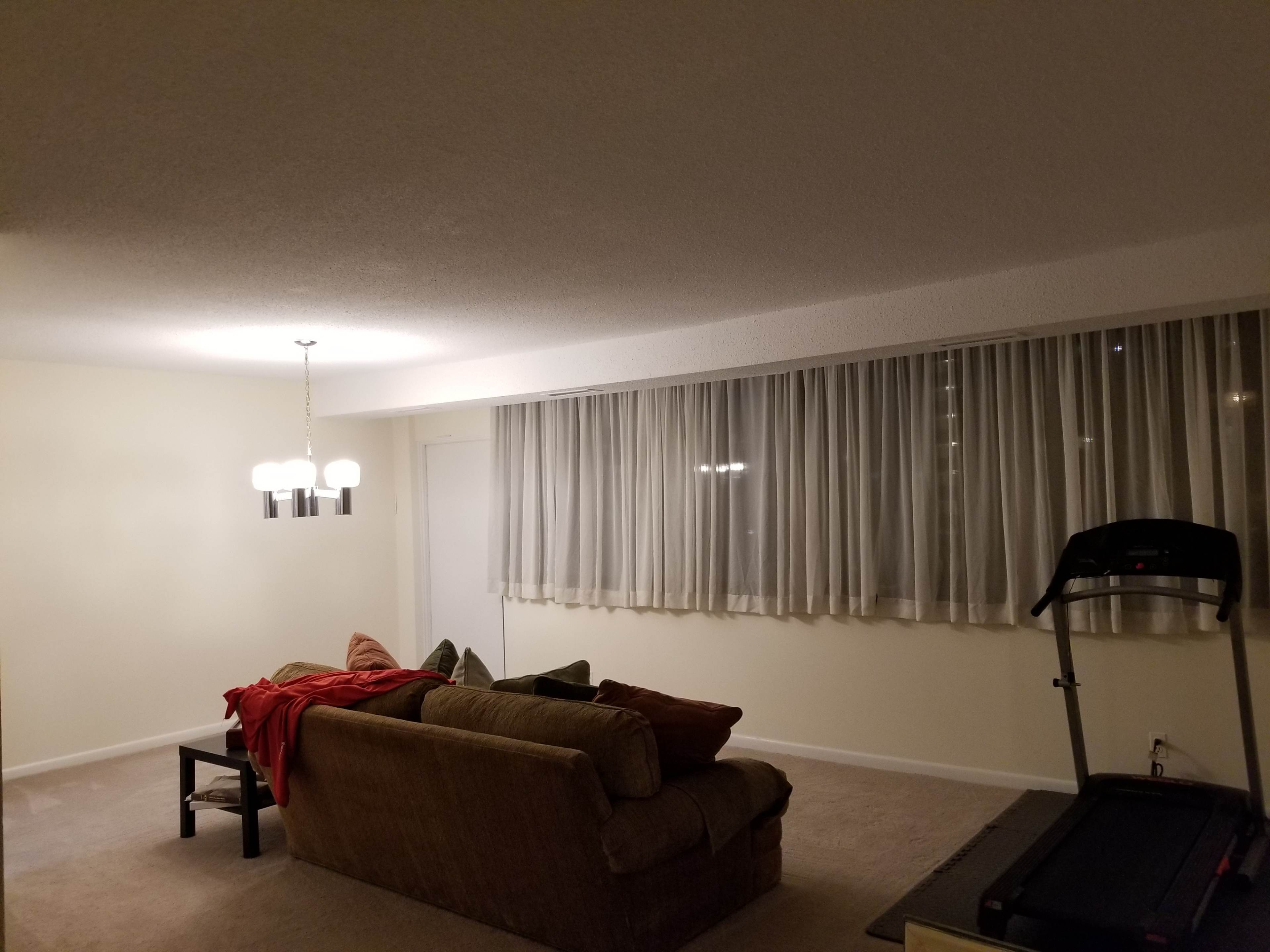 A living room features a couch with a blanket, a treadmill, a chandelier, and large curtains covering a window.