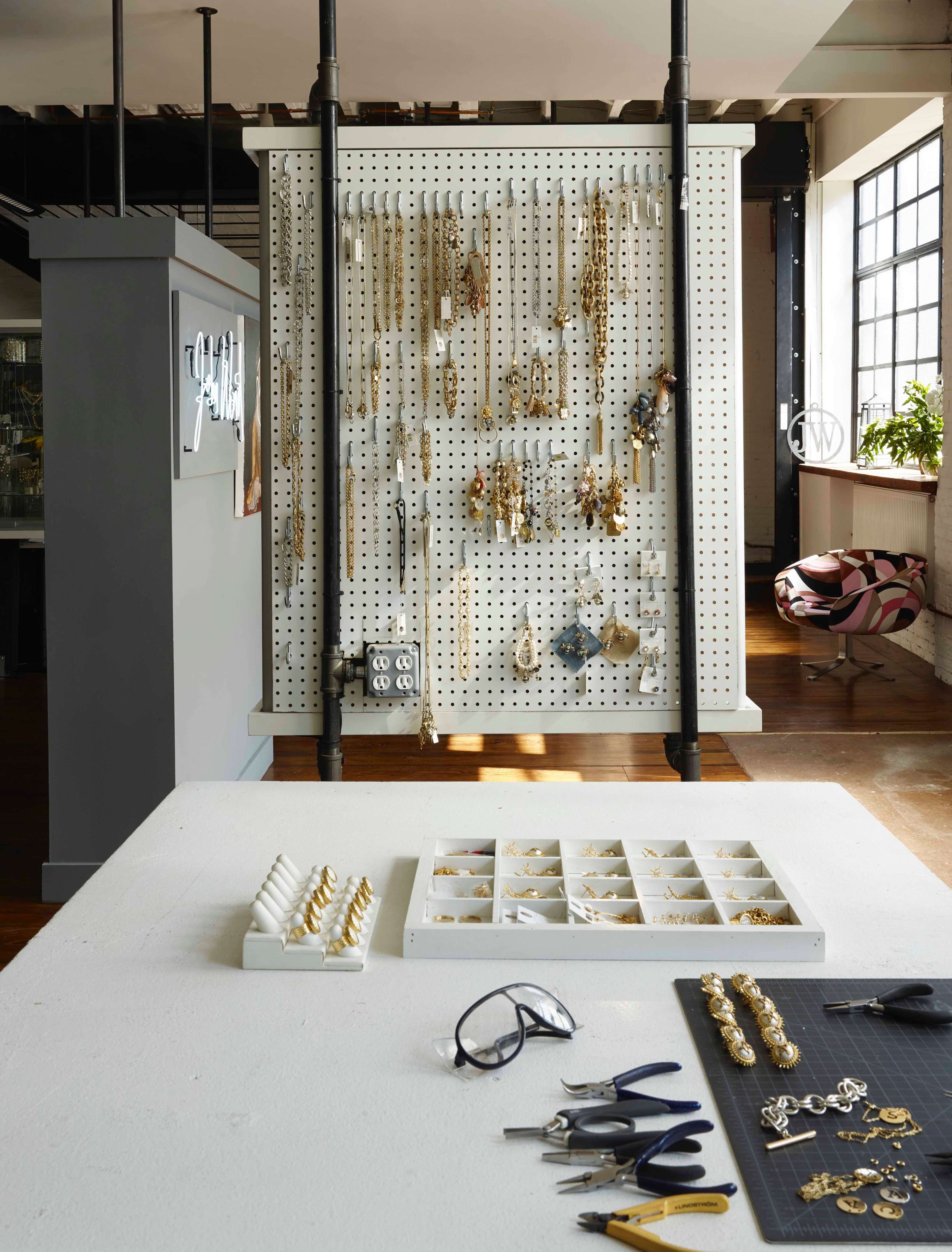 The image shows a workshop setting with a pegboard displaying various necklaces and earrings, while a table holds tools and organized jewelry components.