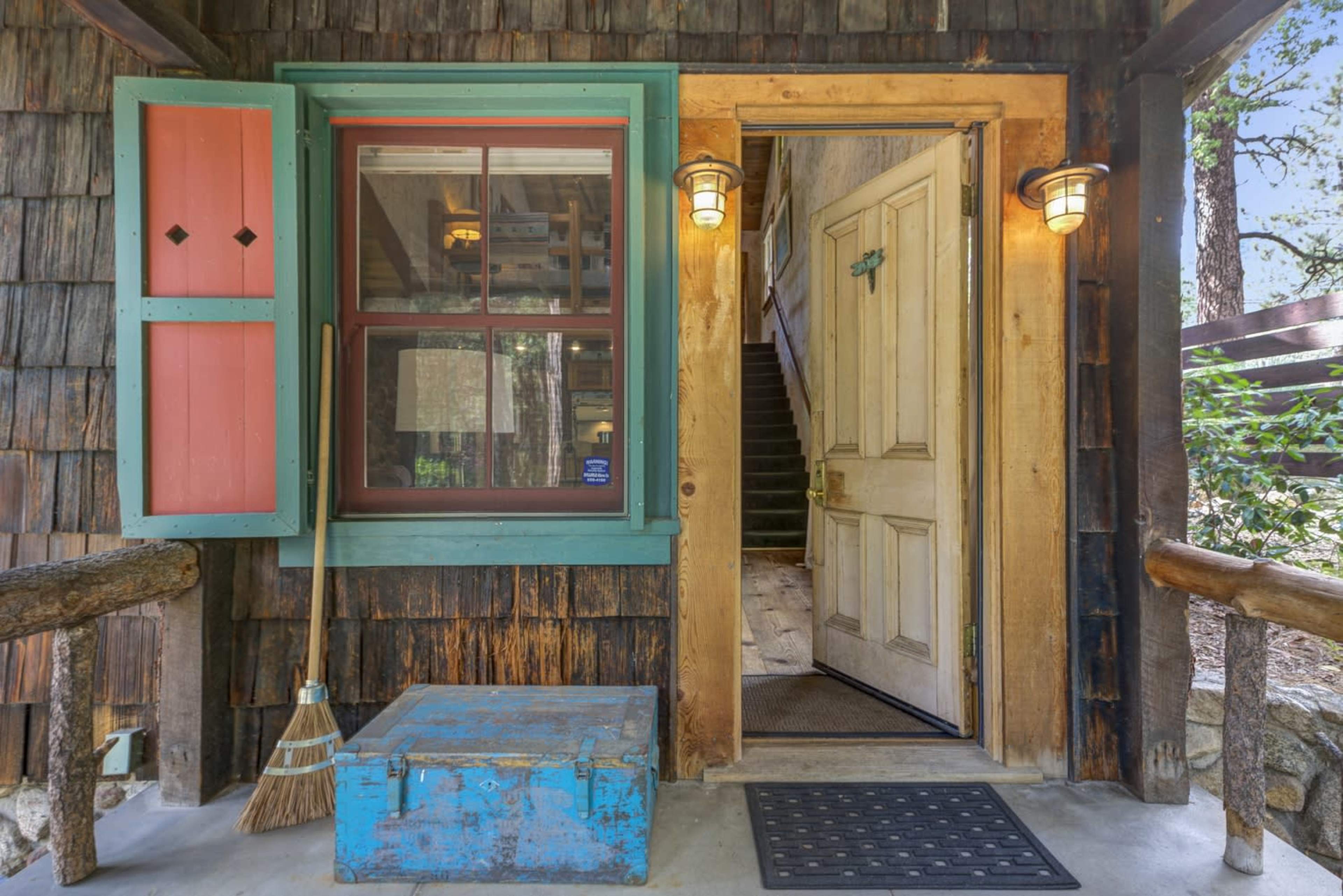 The entrance features a wooden door with colorful trim, flanked by a window with shutters, and a broom beside a blue storage chest.