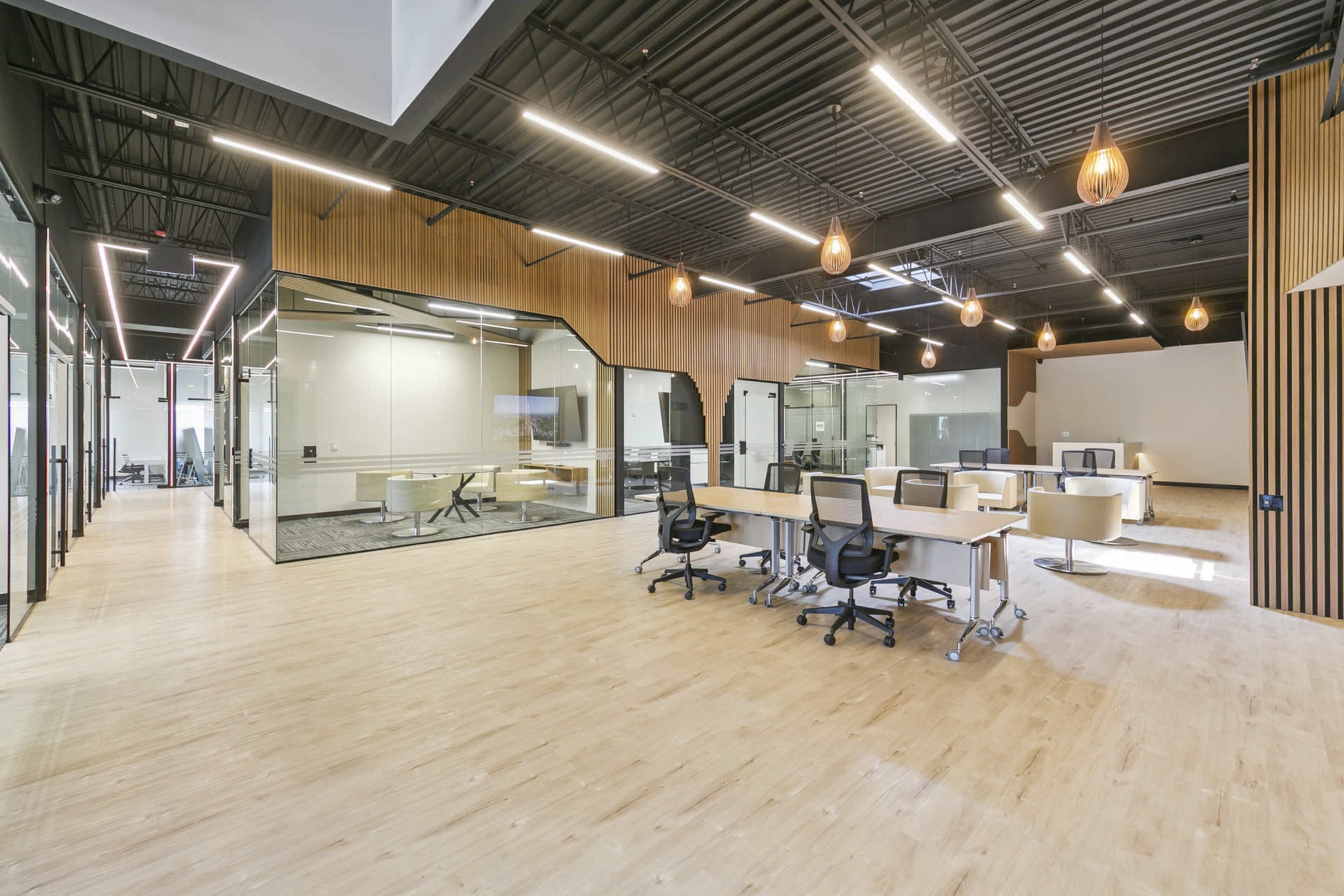 The image shows a modern office space with wooden flooring, large glass meeting rooms, and aesthetic pendant lighting.