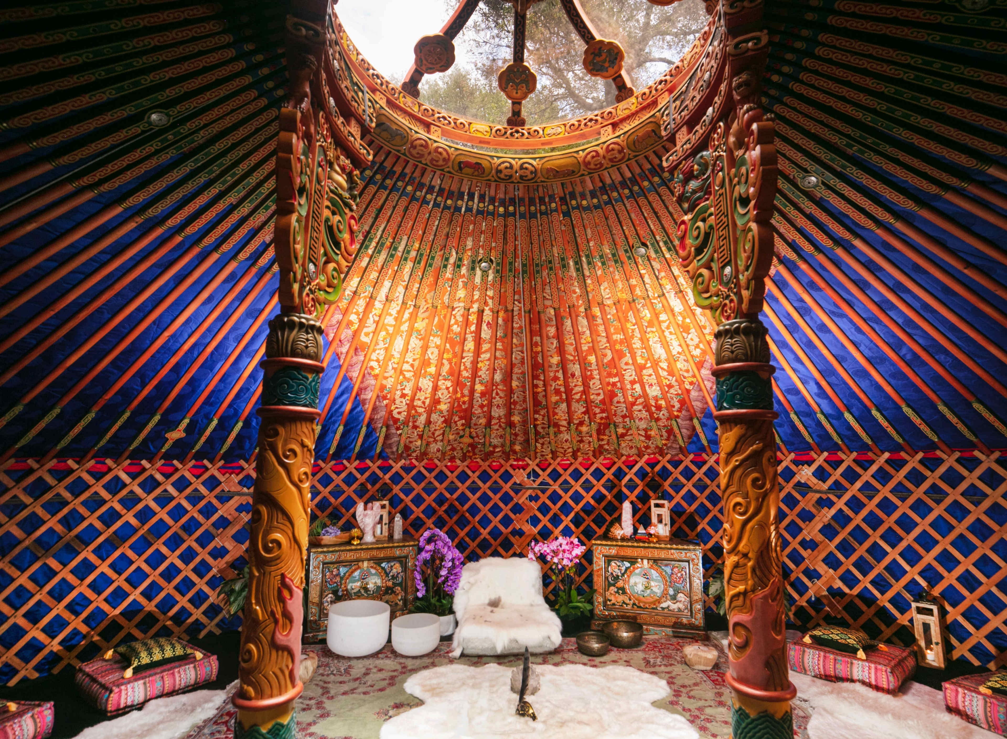 The interior of a yurt features colorful walls adorned with intricate patterns, wooden pillars, and cozy furnishings arranged on a decorative floor covering.