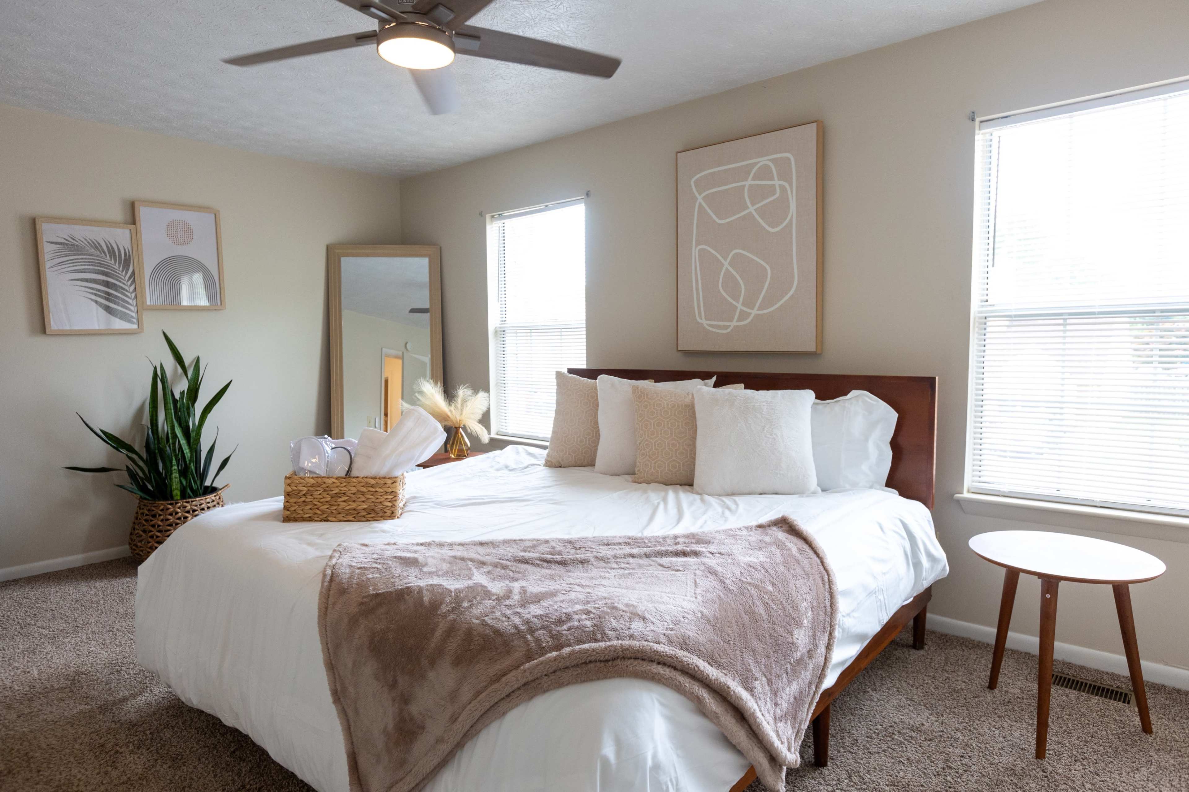 A well-lit bedroom features a large bed with white bedding, a decorative throw, a plant in a corner, and a mirror beside a wooden nightstand.