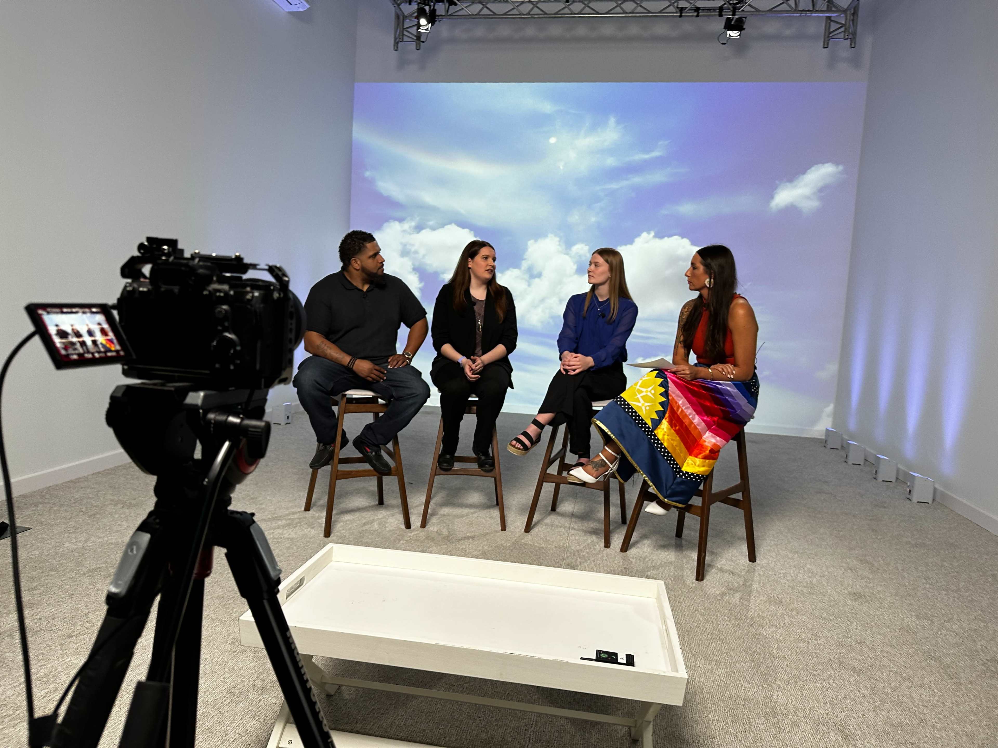 A group of four people sits on stools in front of a large backdrop featuring a cloudy sky, with a camera set up to capture the discussion.